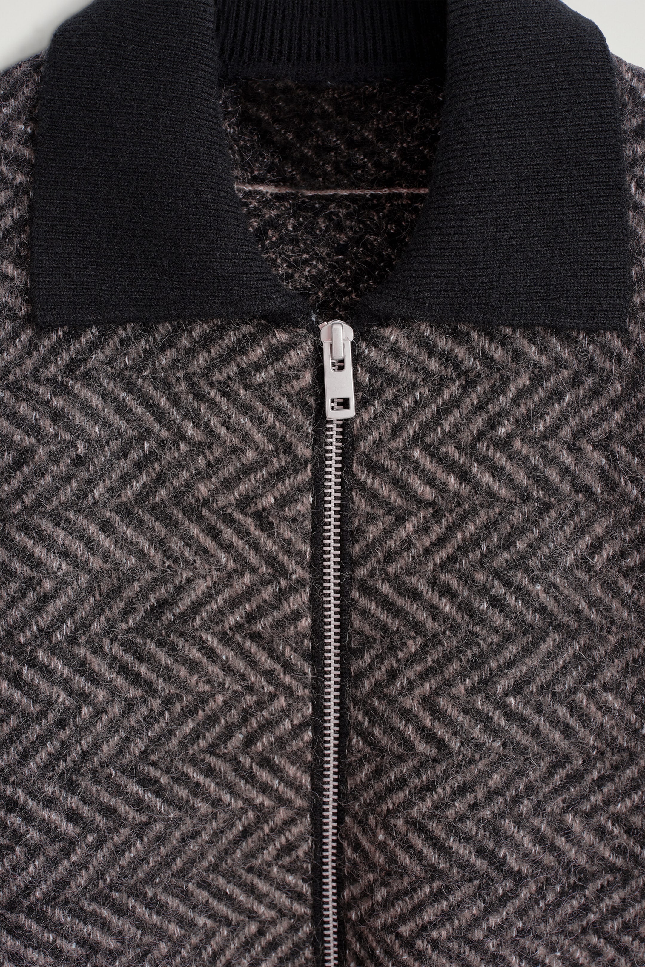 Cette vue rapprochée de face de la veste tricotée met en valeur son motif chevrons texturé gris et noir, son col côtelé noir uni et sa fermeture éclair argentée au centre.