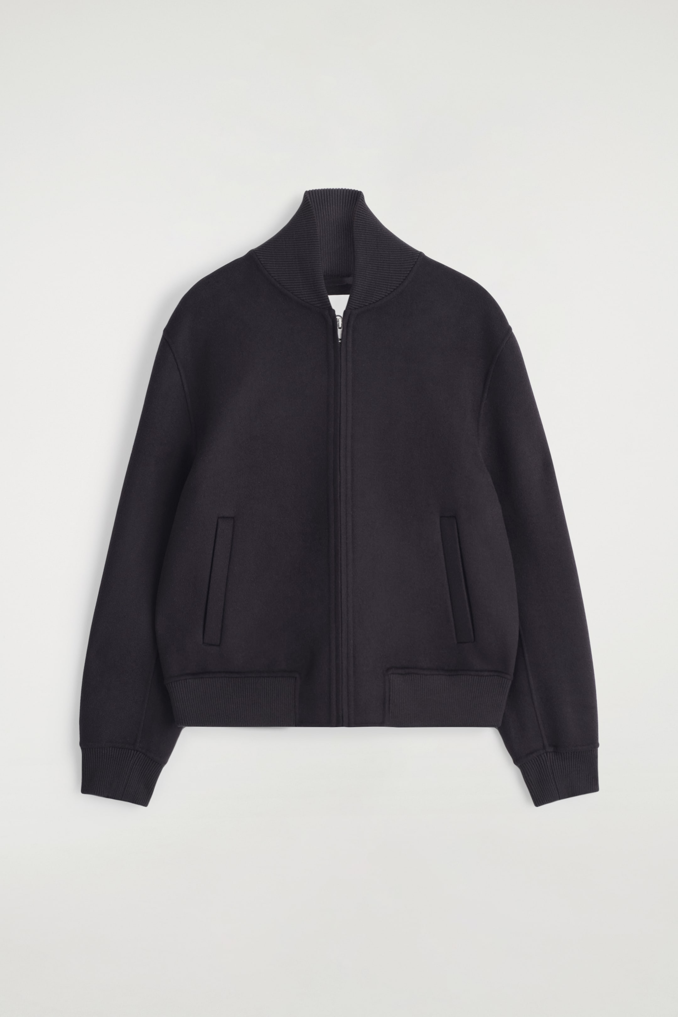 View larger image: A dark-colored bomber jacket is being presented from the front, featuring a full-length zipper closure and a ribbed stand-up collar. Two rectangular welt pockets, ribbed cuffs, and a ribbed hem are also visible on the jacket.