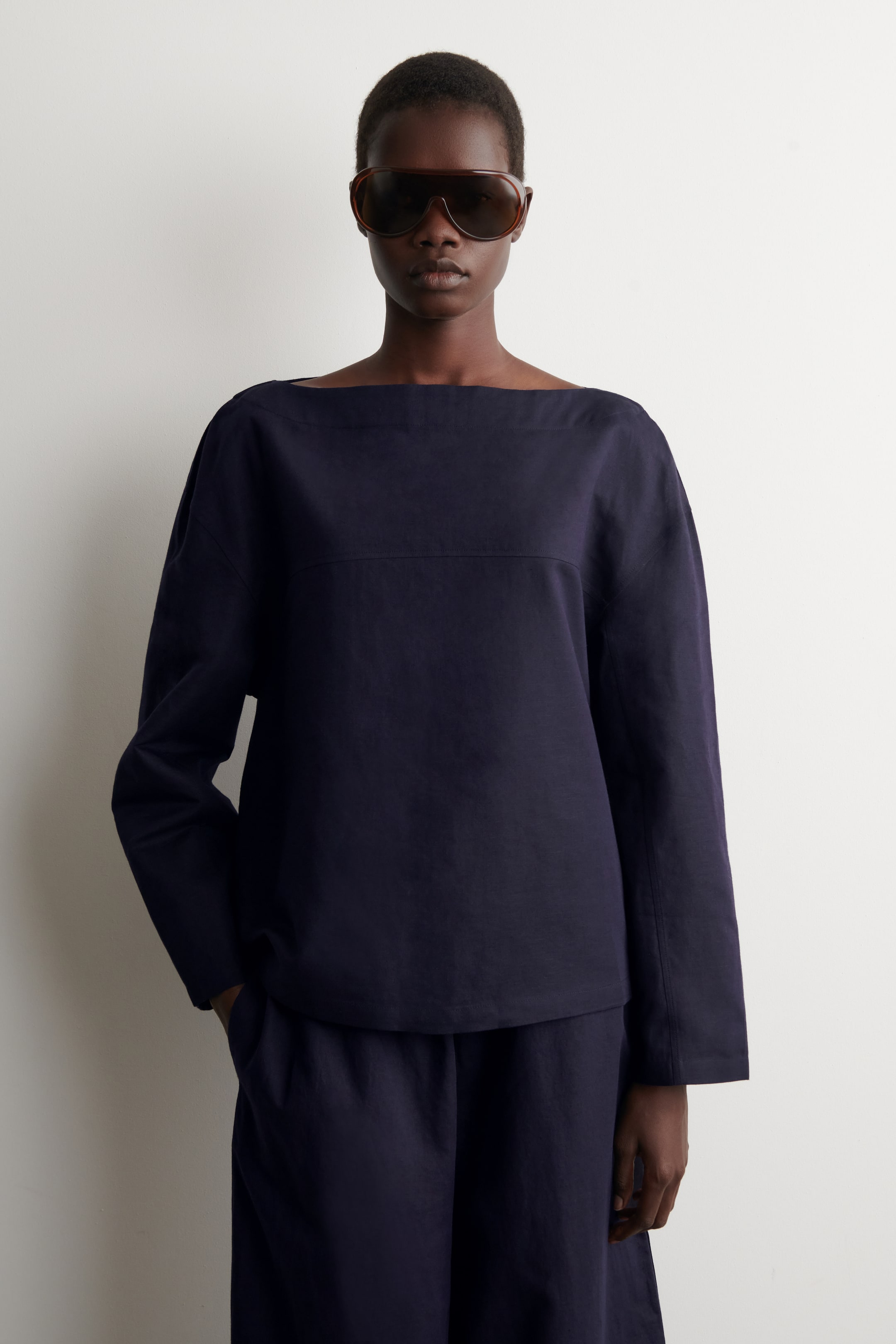 Visualizza l'immagine ingrandita: Una maglia blu scuro con scollo a barchetta presenta maniche lunghe e ampie e una cucitura orizzontale sul petto superiore. È abbinata a pantaloni larghi coordinati, con una mano che riposa in una tasca dei pantaloni.