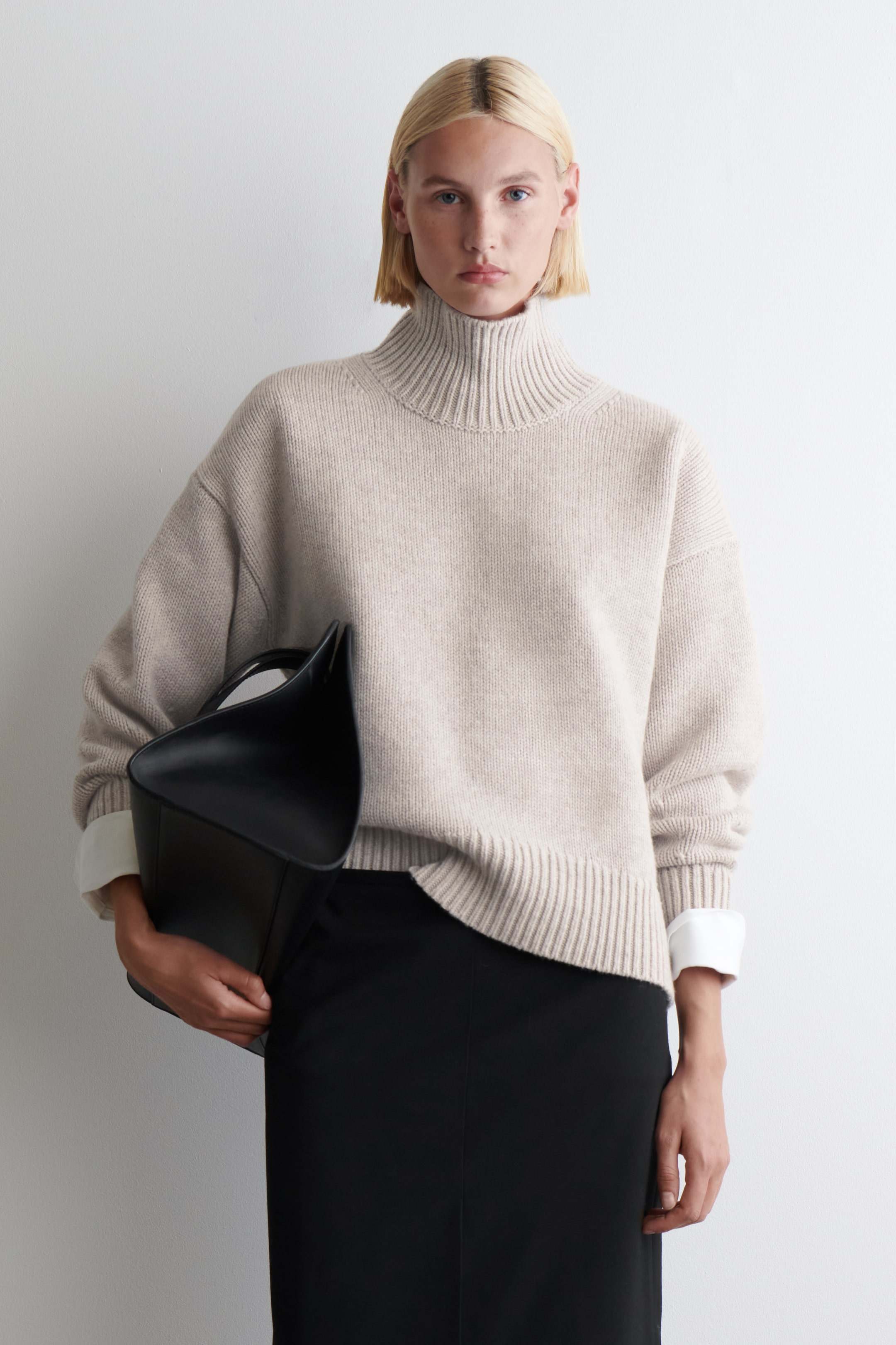 Grösseres Bild ansehen: Ein hellbeiger Rollkragenpullover wird von vorne präsentiert, mit einem gerippten Rollkragen, einer lockeren Passform und gerippten Bündchen und Saum. Weiße Hemdmanschetten liegen darunter und ragen über die Bündchen des Pullovers hinaus.