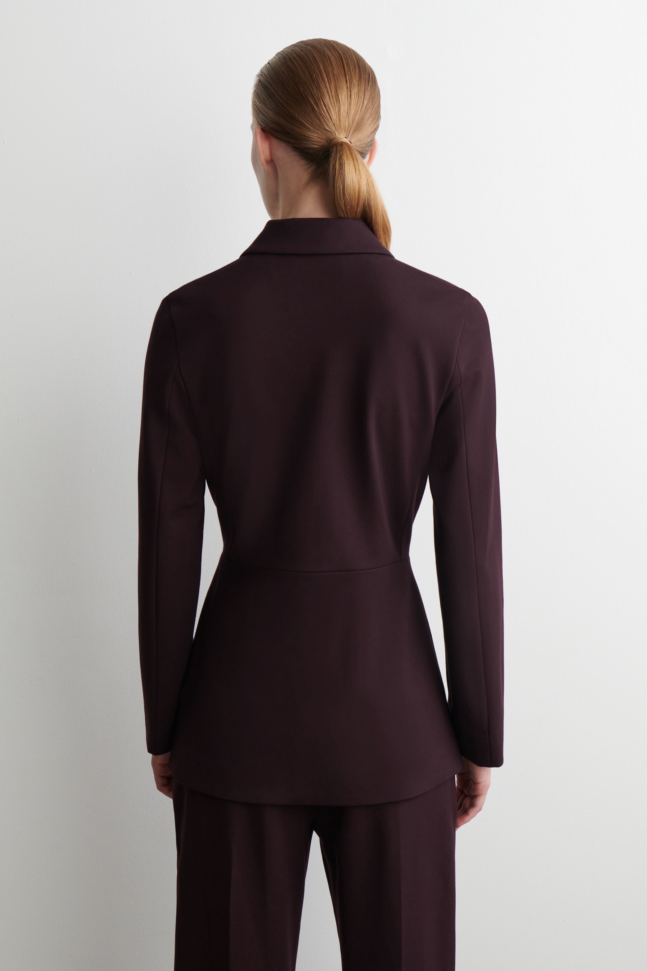 Visualizza immagine più grande: Un blazer color Bordeaux viene presentato dalla vista posteriore, mettendo in mostra la sua silhouette su misura con una cucitura in vita definita. Il tessuto liscio crea un drappeggio fluido, che si estende in maniche a lunghezza intera e un leggero svasamento sotto la vita, ed è indossato con pantaloni coordinati.