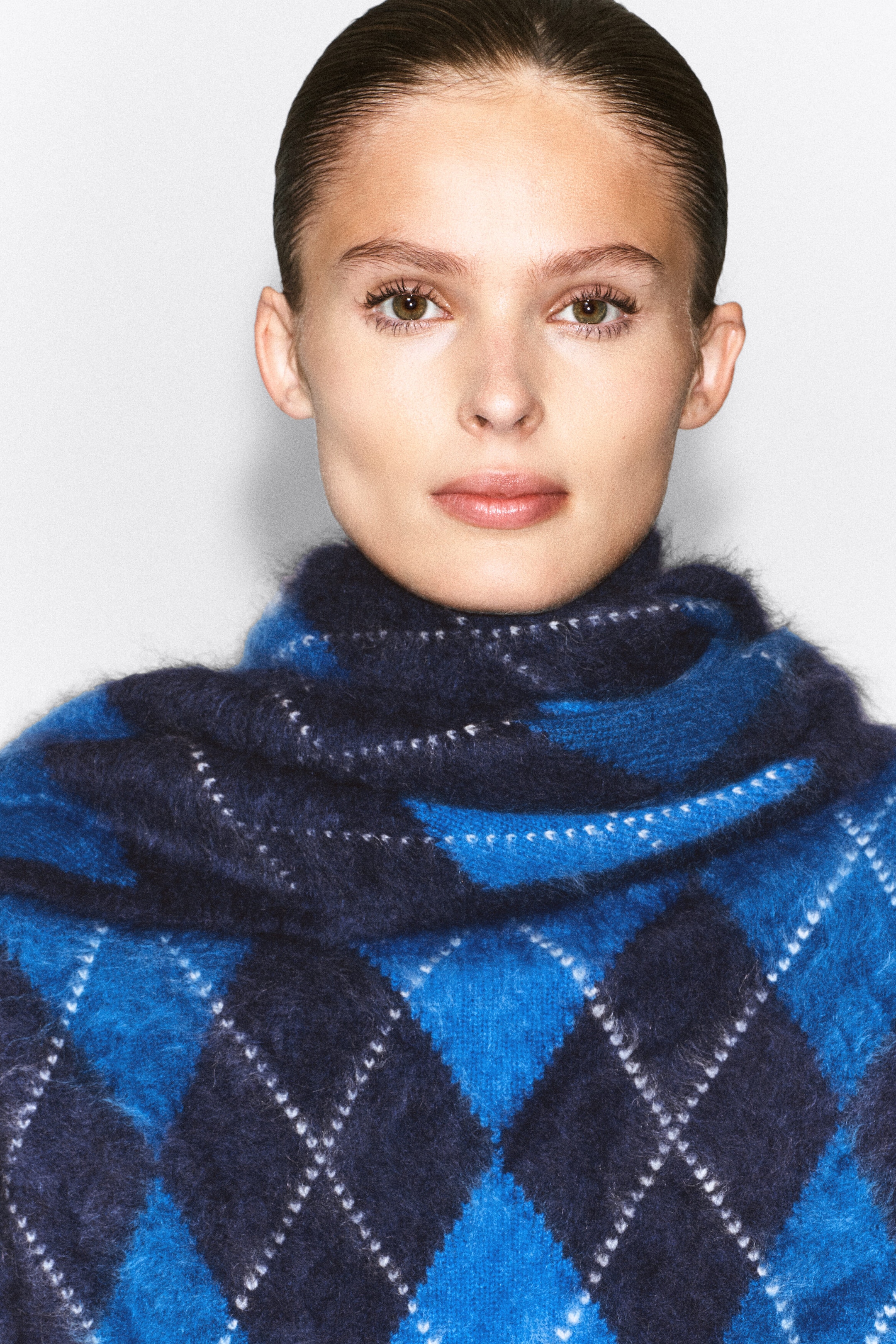An argyle brushed cashmere triangle scarf in shades of blue and navy is worn wrapped around the model’s neck. The scarf features a fuzzy, textured knit with a classic diamond pattern highlighted by white dashed outlines. An argyle brushed cashmere triangle scarf in shades of blue and navy is worn wrapped around the model’s neck. The scarf features a fuzzy, textured knit with a classic diamond pattern highlighted by white dashed outlines.