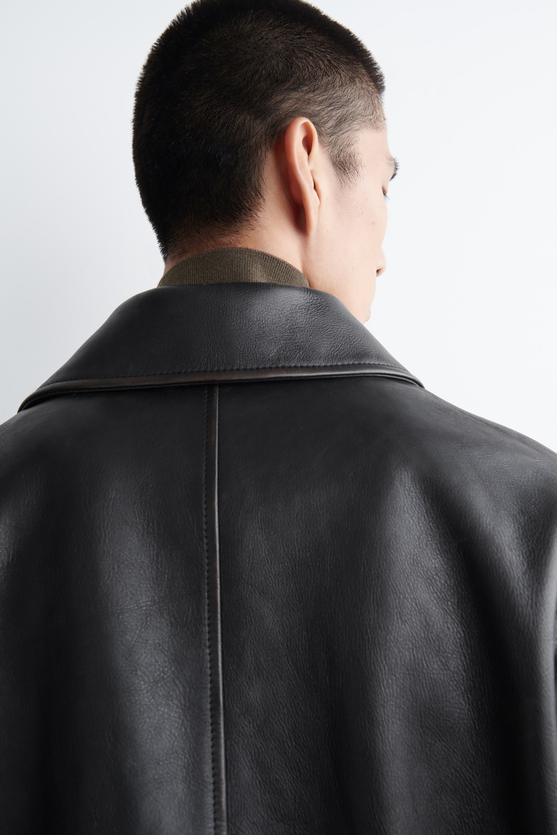 Agrandir l'image: Une veste en cuir noir est montrée de dos, légèrement inclinée vers la gauche. Elle présente un col large et plat et une couture verticale distincte qui descend au centre du dos, avec un col côtelé vert olive foncé visible en dessous.