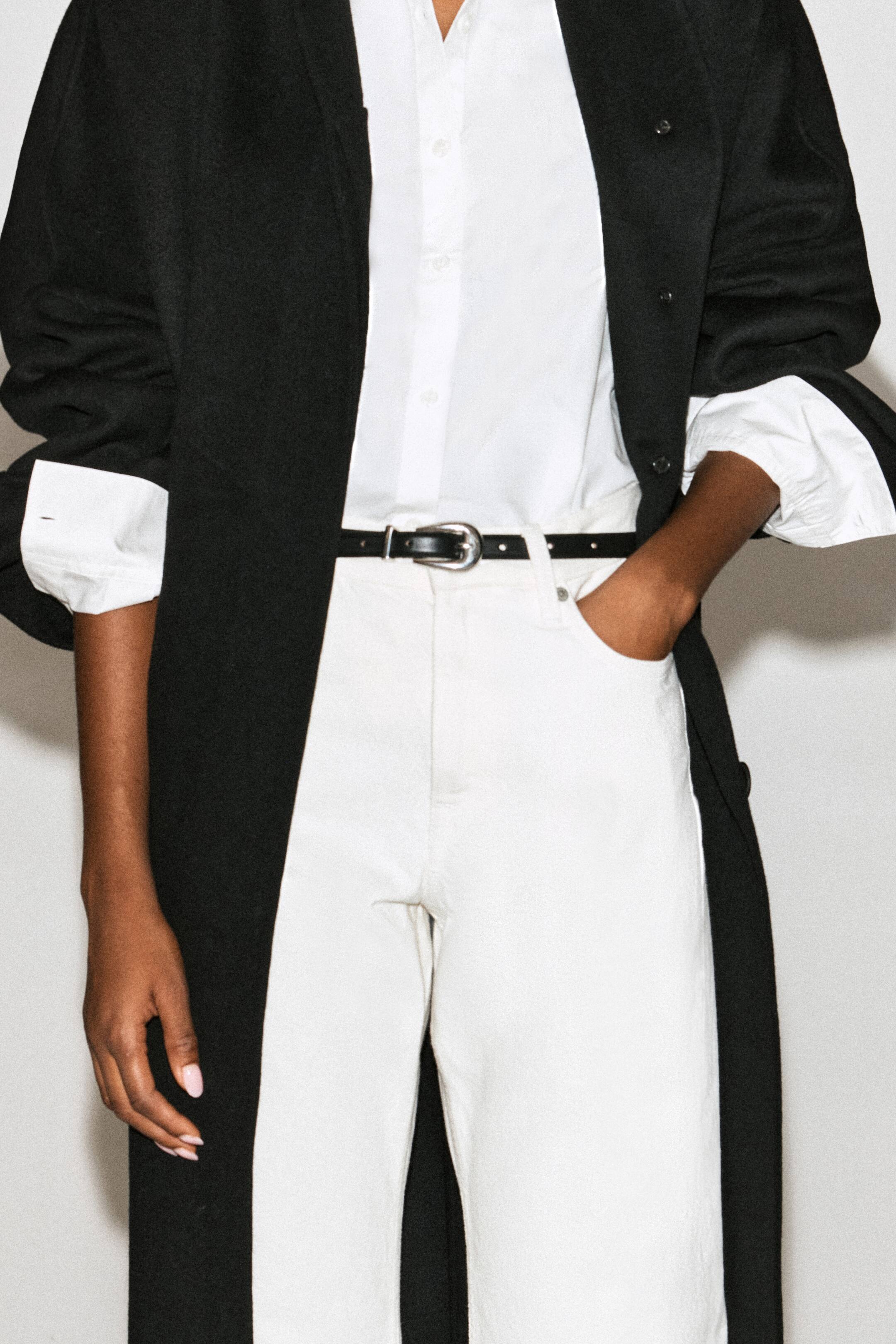 Visualizza l'immagine ingrandita: Una sottile cintura nera in pelle con fibbia scolpita color argento è indossata attraverso i passanti di pantaloni bianchi a vita alta dalla modella. La cintura è abbinata a una camicia bianca abbottonata infilata nei pantaloni e indossata sotto un lungo cappotto nero.
