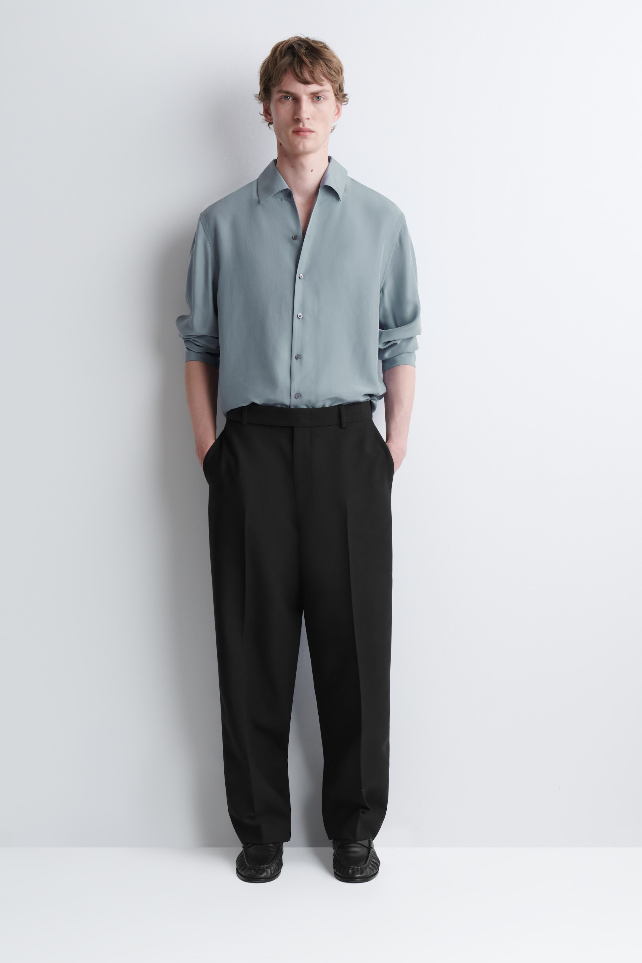Agrandir l'image: Un pantalon droit en mélange de laine noir est porté par le mannequin, qui se tient debout les mains posées dans les poches latérales. Le pantalon présente des plis marqués le long de l’avant des jambes et est associé à une chemise à manches longues bleu clair rentrée dans la ceinture.