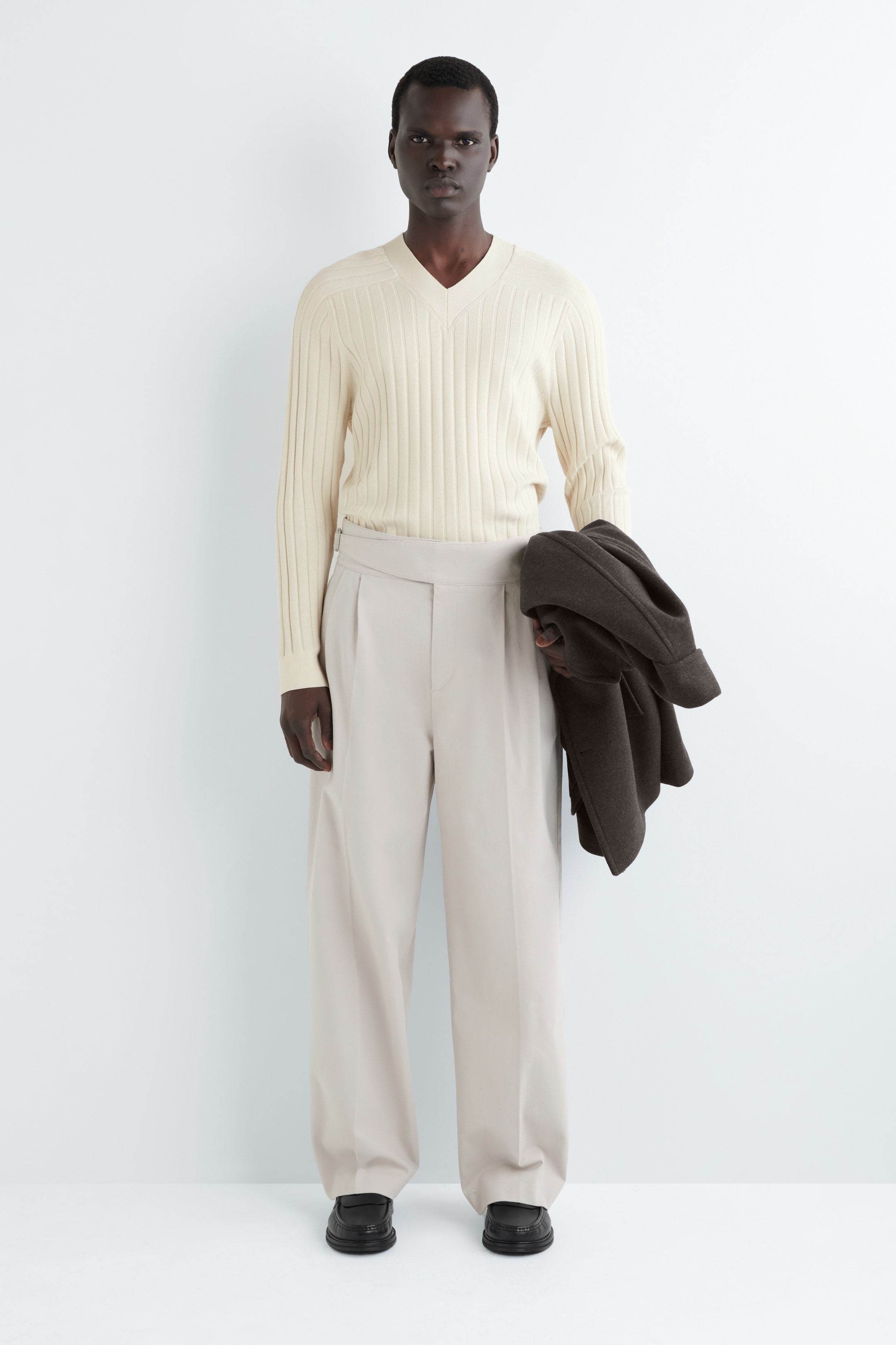 Agrandir l'image: Un pull crème à col en V, avec des côtes verticales, est porté avec un pantalon large plissé beige clair et des mocassins noirs épais, tous tournés vers l'avant. Une veste marron foncé est tenue au-dessus du bras gauche.