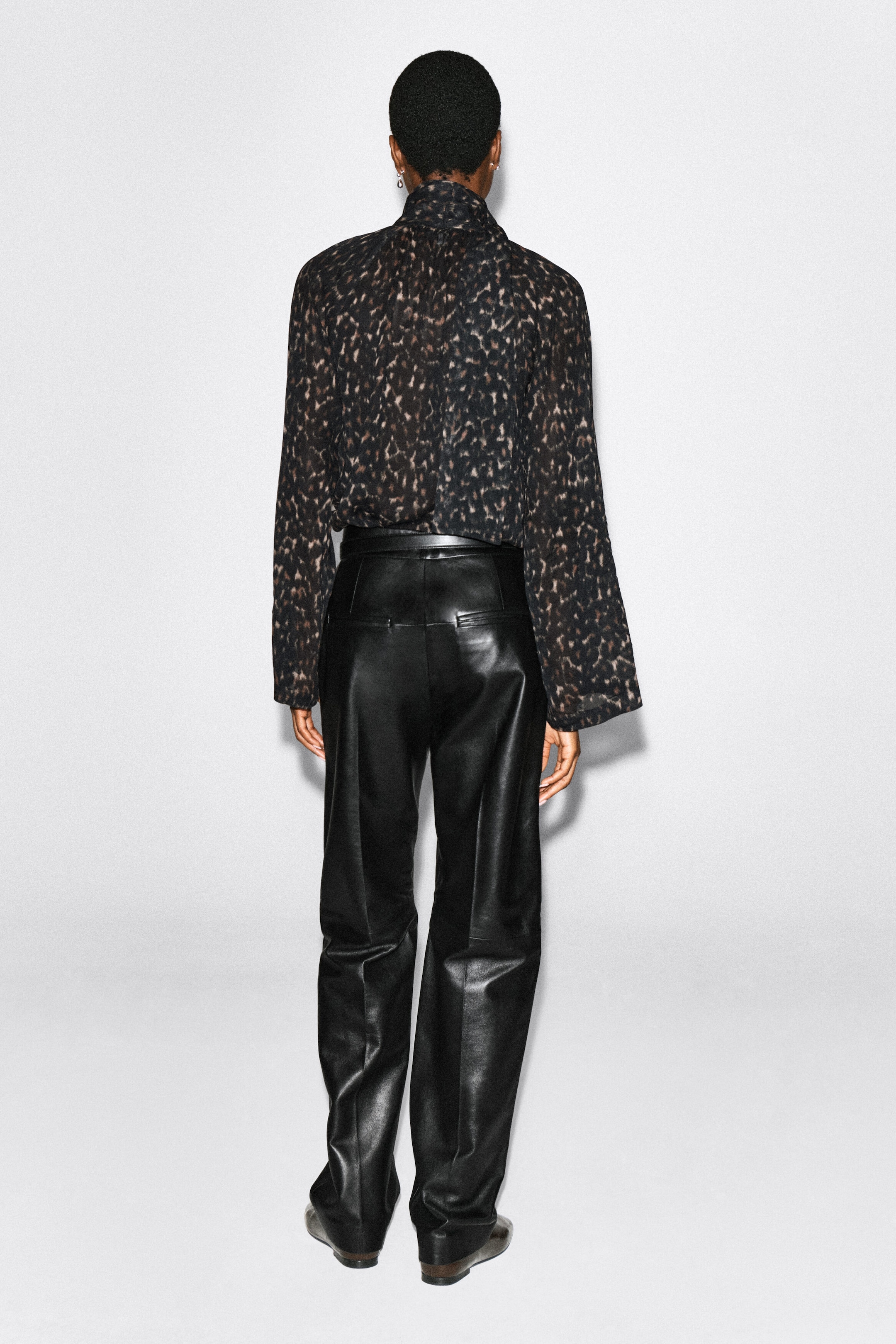 A leopard-print fluid scarf-detail top is being worn by the model, viewed from the back. This semi-sheer, long-sleeved top features a high neckline and is tucked into a pair of black leather trousers. A leopard-print fluid scarf-detail top is being worn by the model, viewed from the back. This semi-sheer, long-sleeved top features a high neckline and is tucked into a pair of black leather trousers.