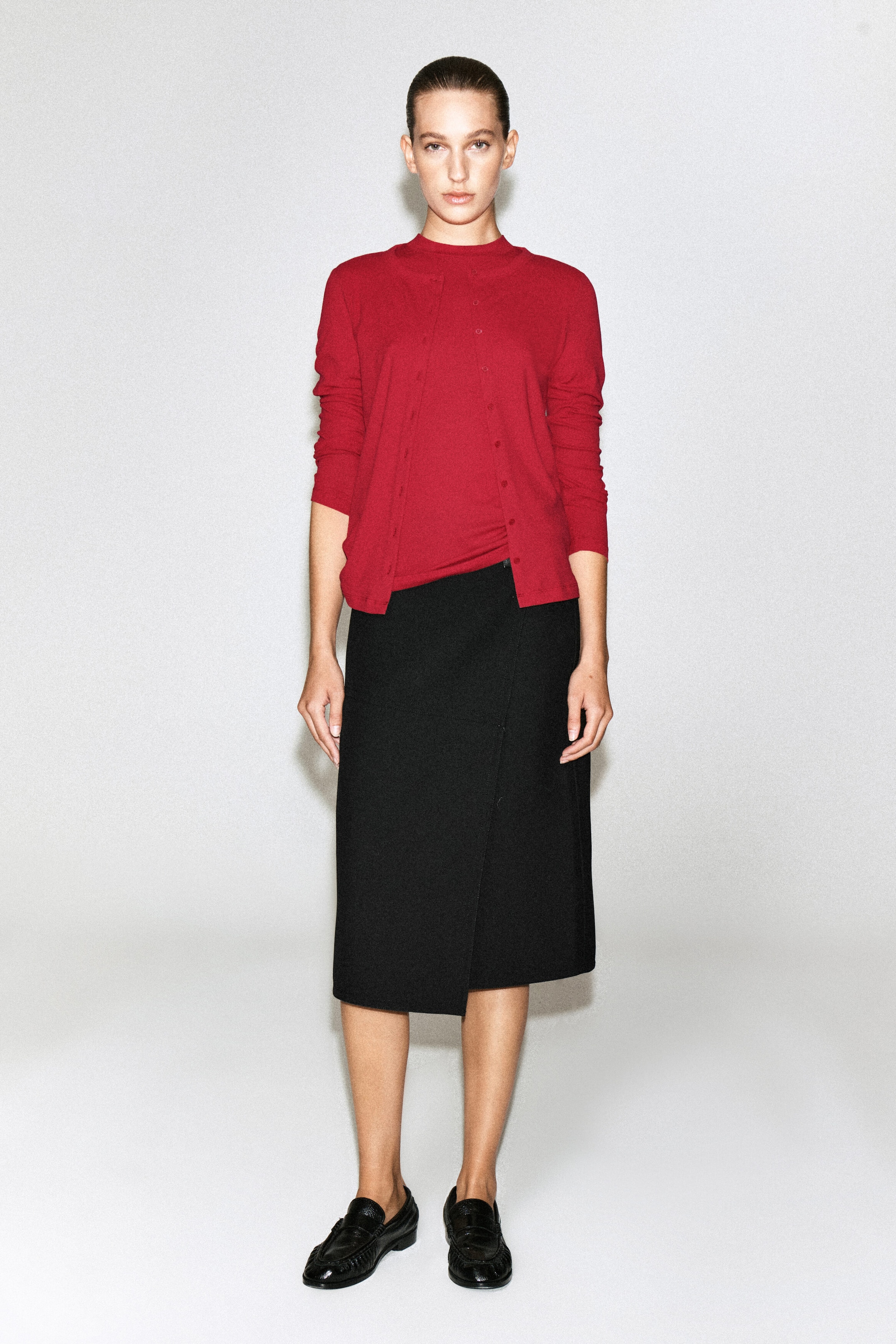 A red jersey cardigan top featuring a button-front closure and long sleeves is being worn by the model, layered over a matching red crew-neck top. The knitwear is styled with a black midi wrap skirt and black loafers. A red jersey cardigan top featuring a button-front closure and long sleeves is being worn by the model, layered over a matching red crew-neck top. The knitwear is styled with a black midi wrap skirt and black loafers.