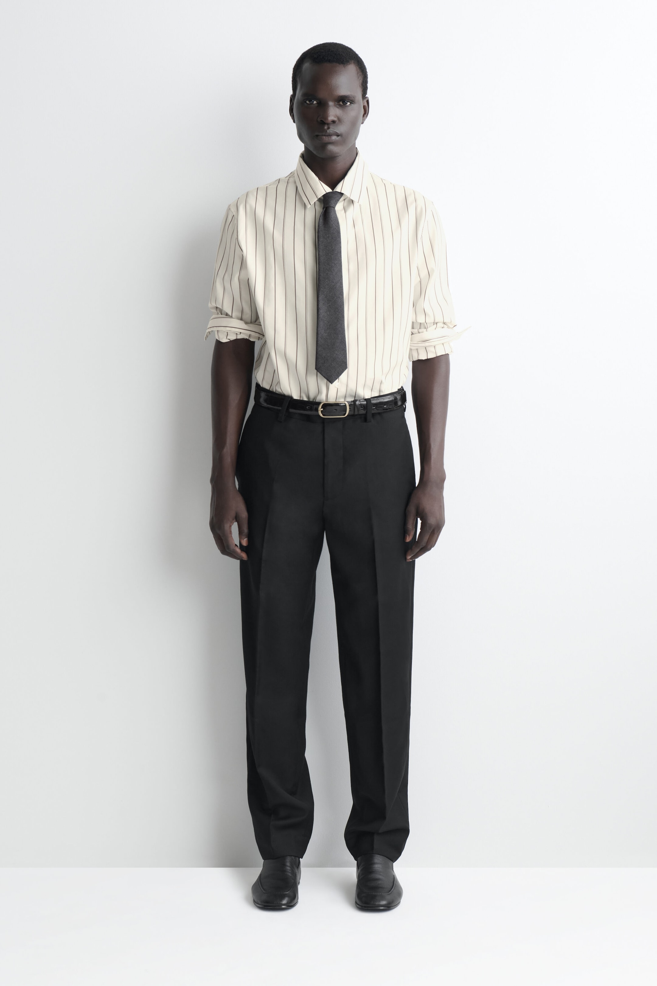 Presented in a front-facing view, a cream-coloured, vertically striped dress shirt with rolled-up sleeves is worn tucked into black, straight-leg trousers with sharp centre creases. The shirt is styled with a dark grey textured tie, and the trousers are secured with a black leather belt. Presented in a front-facing view, a cream-coloured, vertically striped dress shirt with rolled-up sleeves is worn tucked into black, straight-leg trousers with sharp centre creases. The shirt is styled with a dark grey textured tie, and the trousers are secured with a black leather belt.