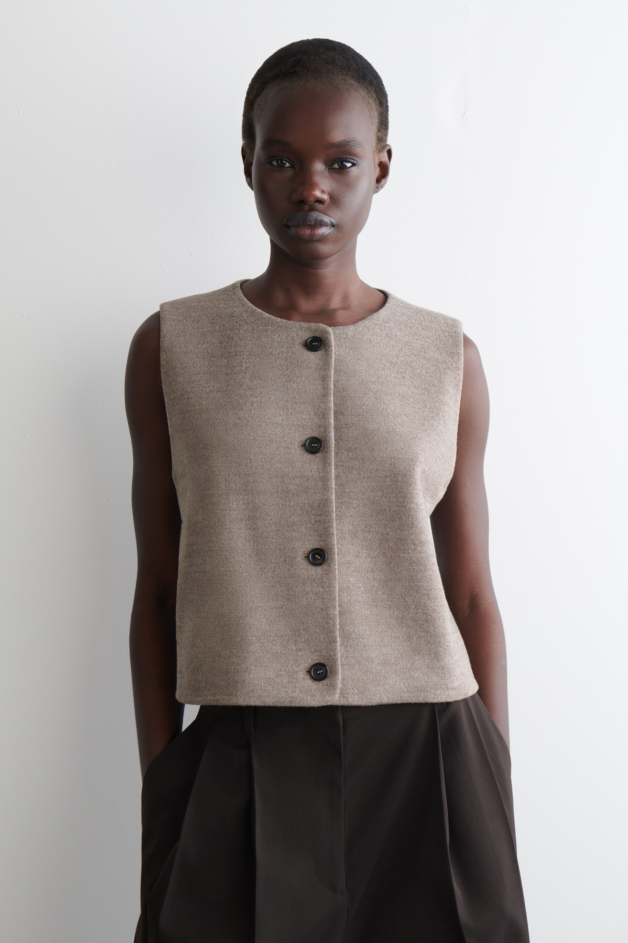 View larger image: A light brown sleeveless waistcoat is being presented from the FRONT, featuring a rounded neckline and a central button closure with four dark buttons. The waistcoat is made from a textured fabric, displaying a regular fit and a cropped length as it is being worn over dark trousers.