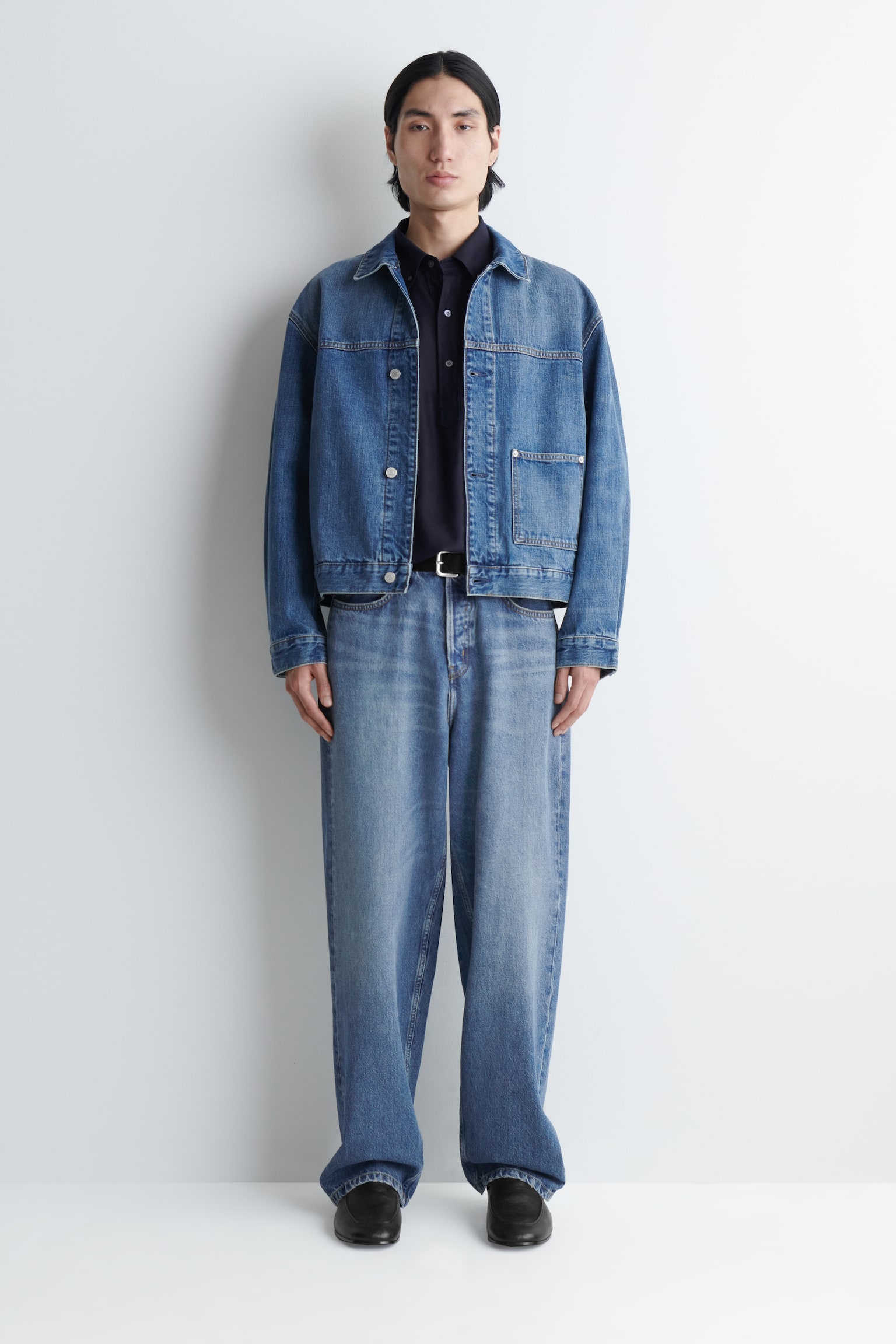 Een blauw denim overhemdjack met een ontspannen pasvorm, zilverkleurige knopen, klinknagels, tabakskleurige stiksels en een enkele opgestikte zak wordt door het model gedragen over een donker shirt. - 1