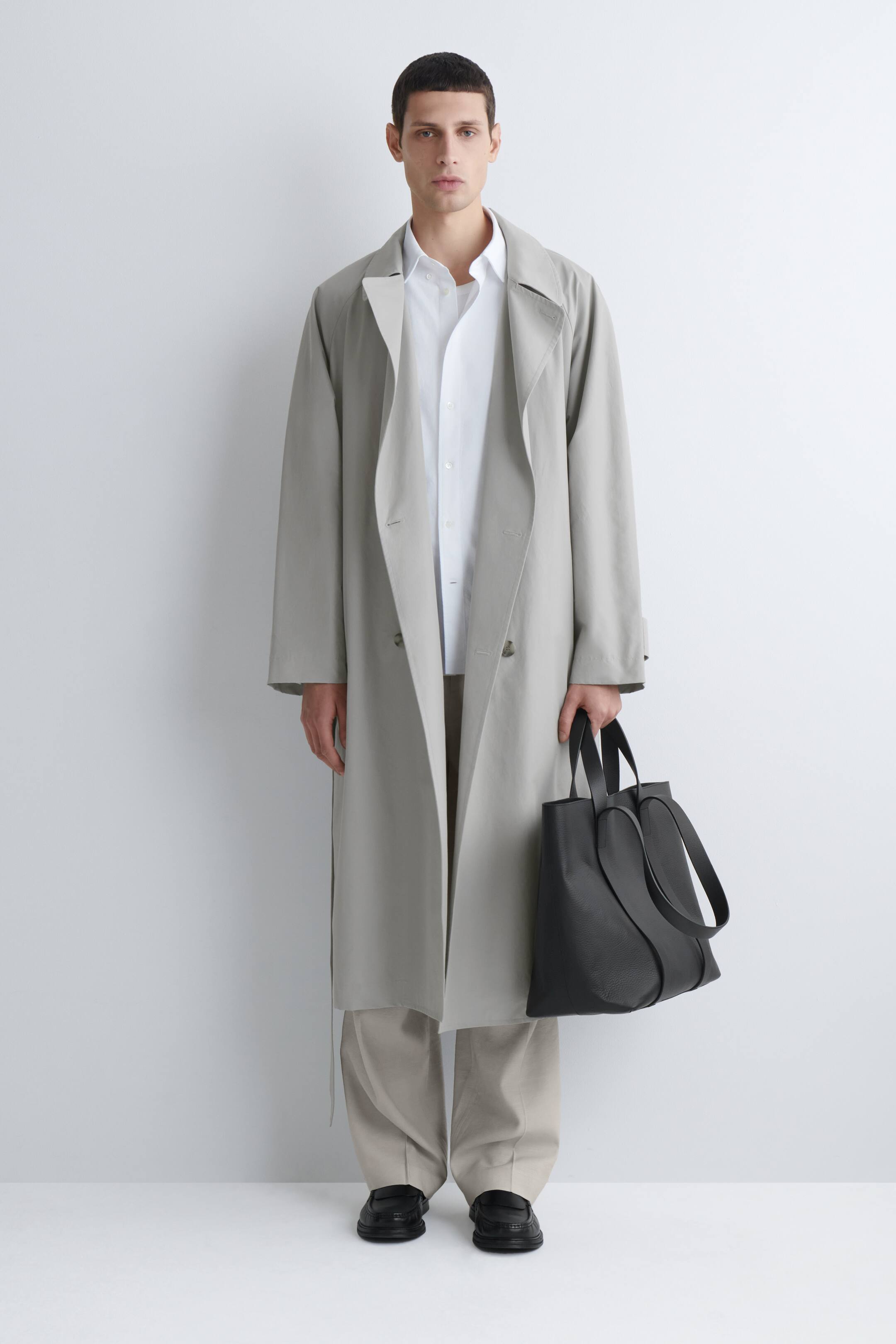הצגת תמונה גדולה יותר: מעיל טרנץ' כפול-שורה בצבע אפור בהיר מוצג מלפנים. הוא כולל דשי ז'קט רחבים בסגנון נוץ' וסילואטה ארוכה ורפויה, הנופלת פתוחה וחושפת חולצה לבנה עם צווארון ומכנסיים בצבע בז' מתחת.