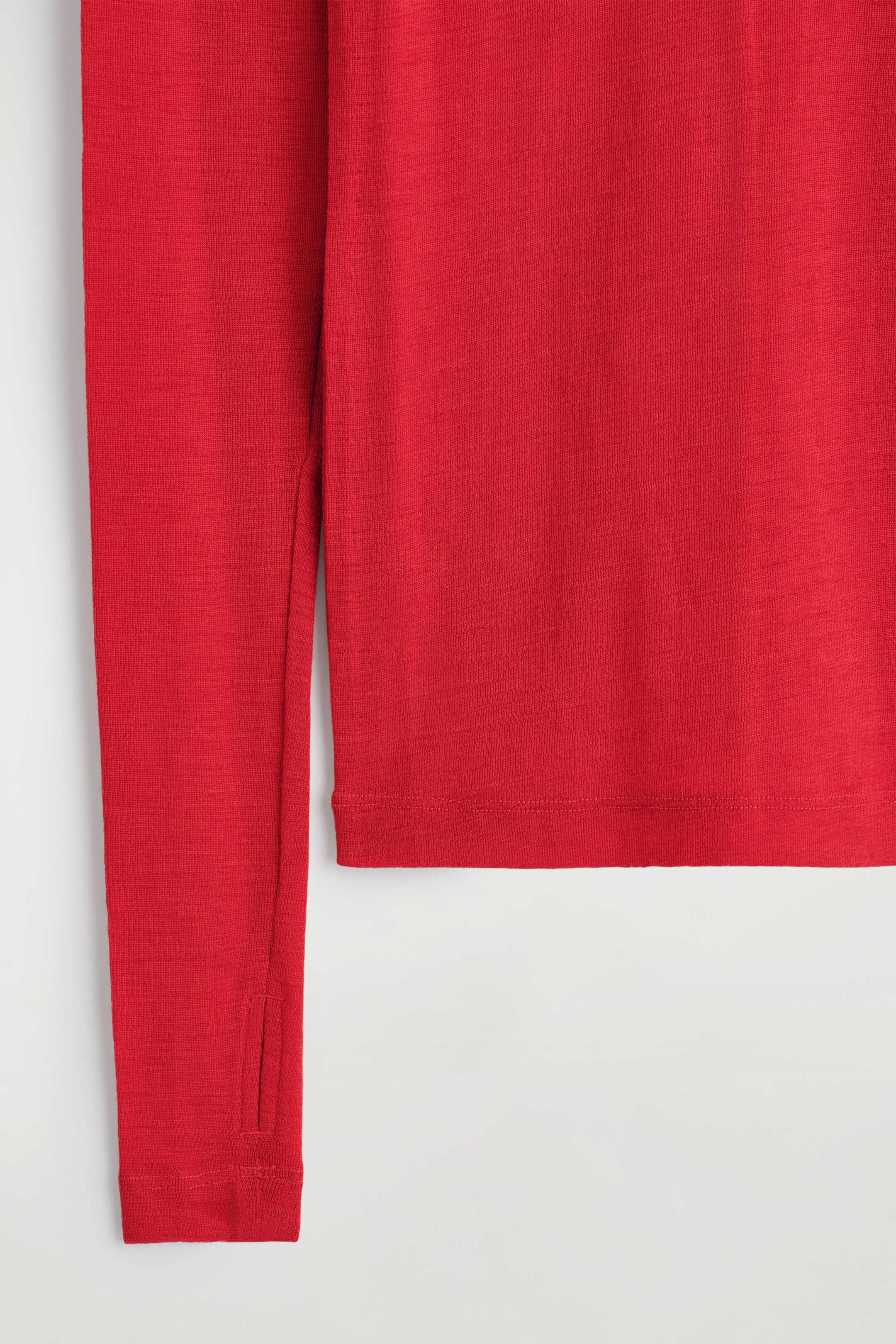 Agrandir l'image: Un haut rouge à manches longues, finement tricoté, expose sa section avant inférieure. Le vêtement présente un ourlet inférieur fini et une manche avec une manchette trou pour le pouce.