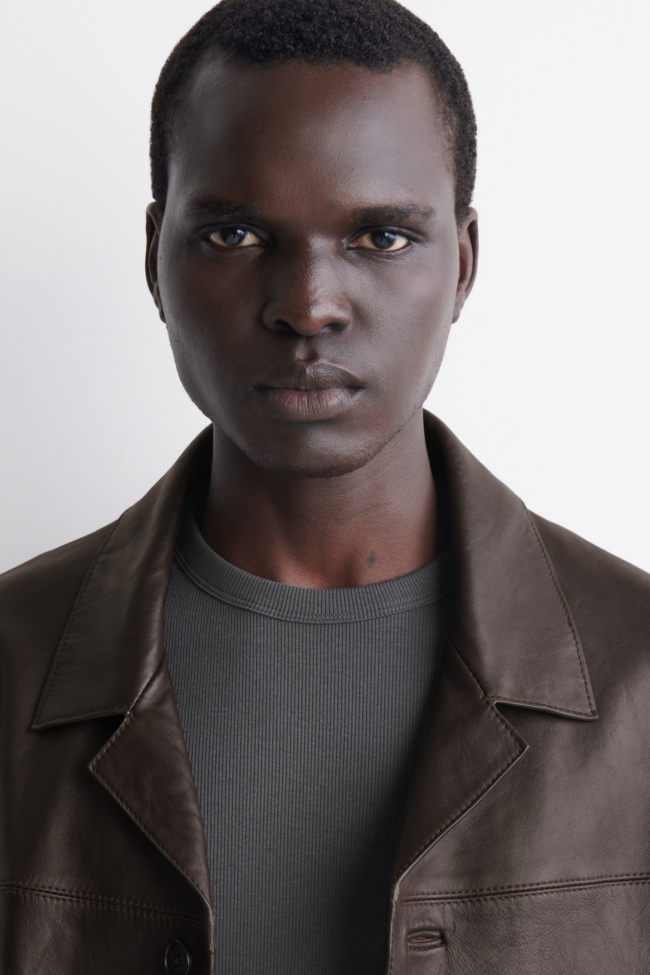 A grey slim ribbed cotton T-shirt is being worn as a base layer under a brown leather jacket, showcasing its fine vertical ribbing and snug fit. This close-up front view highlights the crew neckline and the textured fabric of the top.