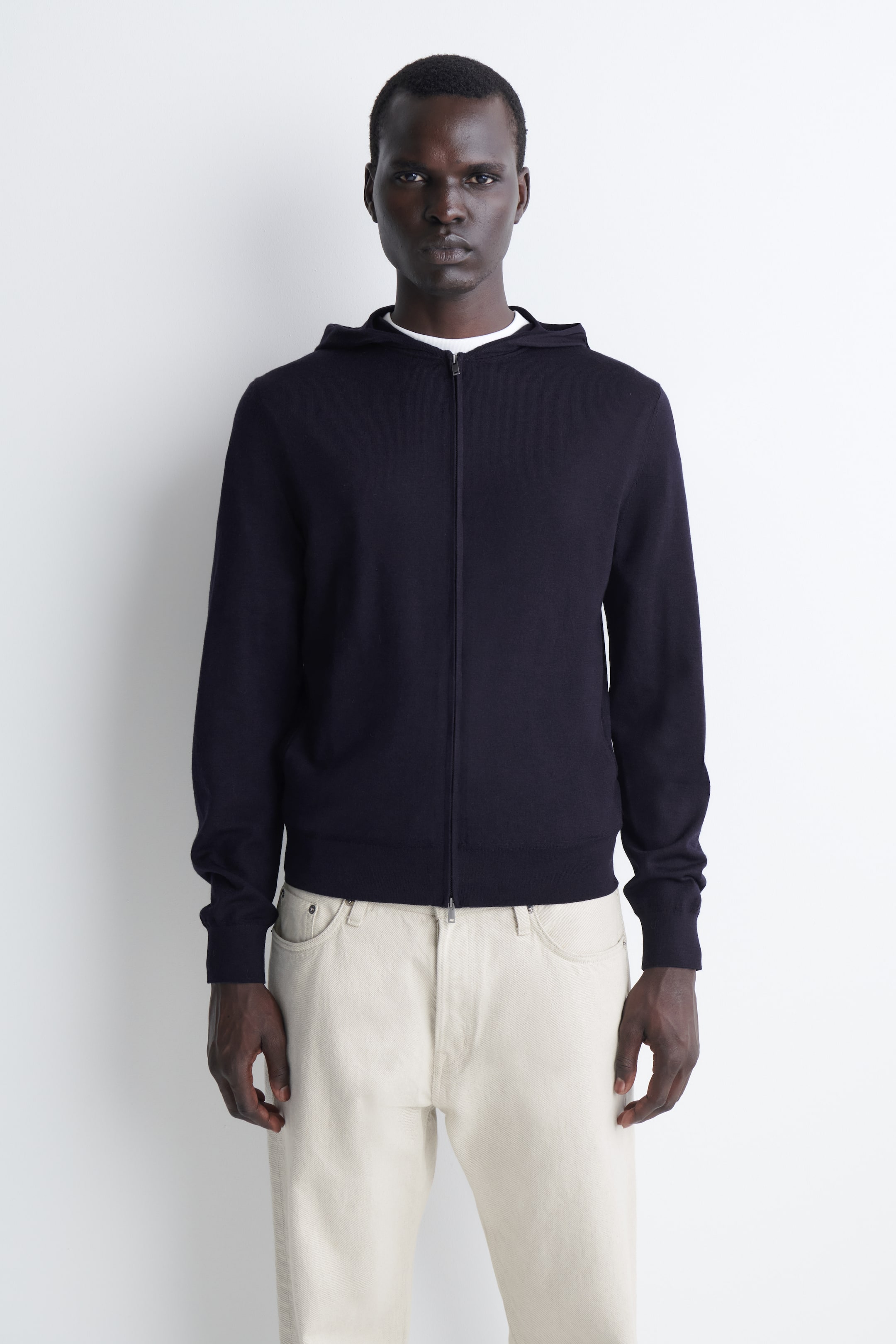 Größeres Bild ansehen: Ein marineblauer Merino-Woll-Hoodie mit Reißverschluss wird in frontaler Ansicht gezeigt und verfügt über eine feinstrukturierte Strickoptik, einen durchgehenden Frontreißverschluss, eine angesetzte Kapuze sowie Rippbündchen an Ärmeln und Saum. Der Hoodie wird über einem weißen T-Shirt mit Rundhalsausschnitt getragen, um seine Slim-Fit-Passform hervorzuheben.