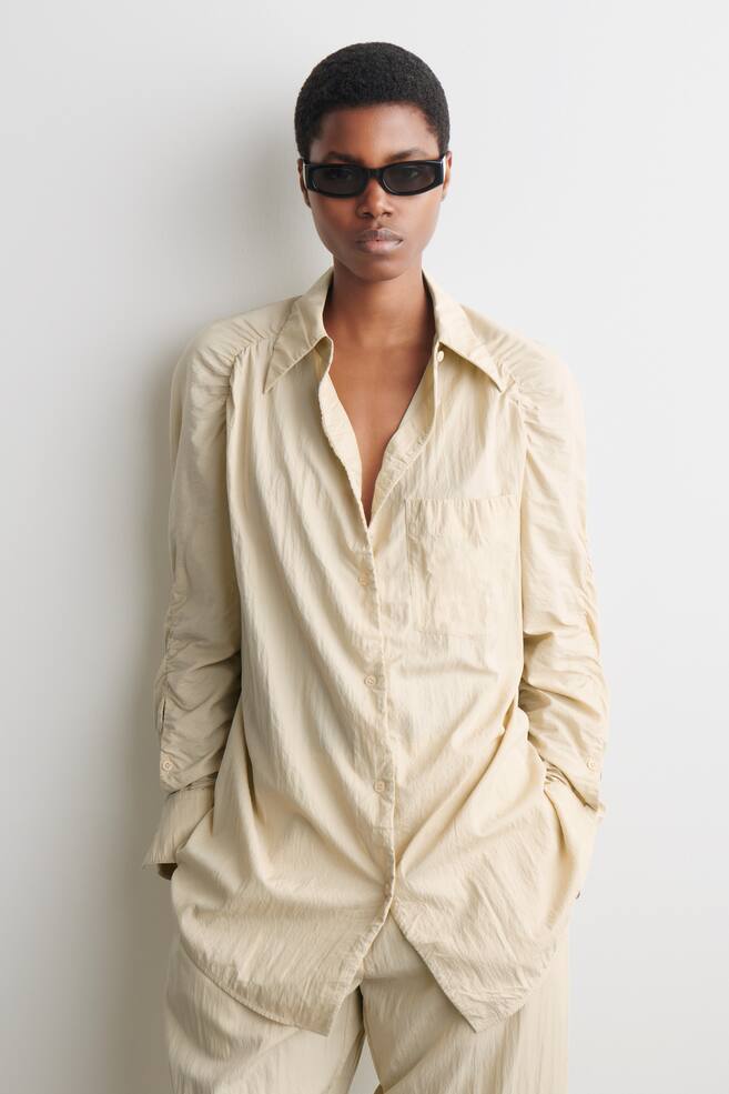 La camisa beige claro está de frente, con un cuello en punta, cierre frontal con botones crema y un único bolsillo en el pecho. Sus mangas largas están recogidas en los hombros y fruncidas, con la manga izquierda aparentemente arremangada. La camisa presenta un ajuste holgado con una textura arrugada y un dobladillo redondeado. - 1