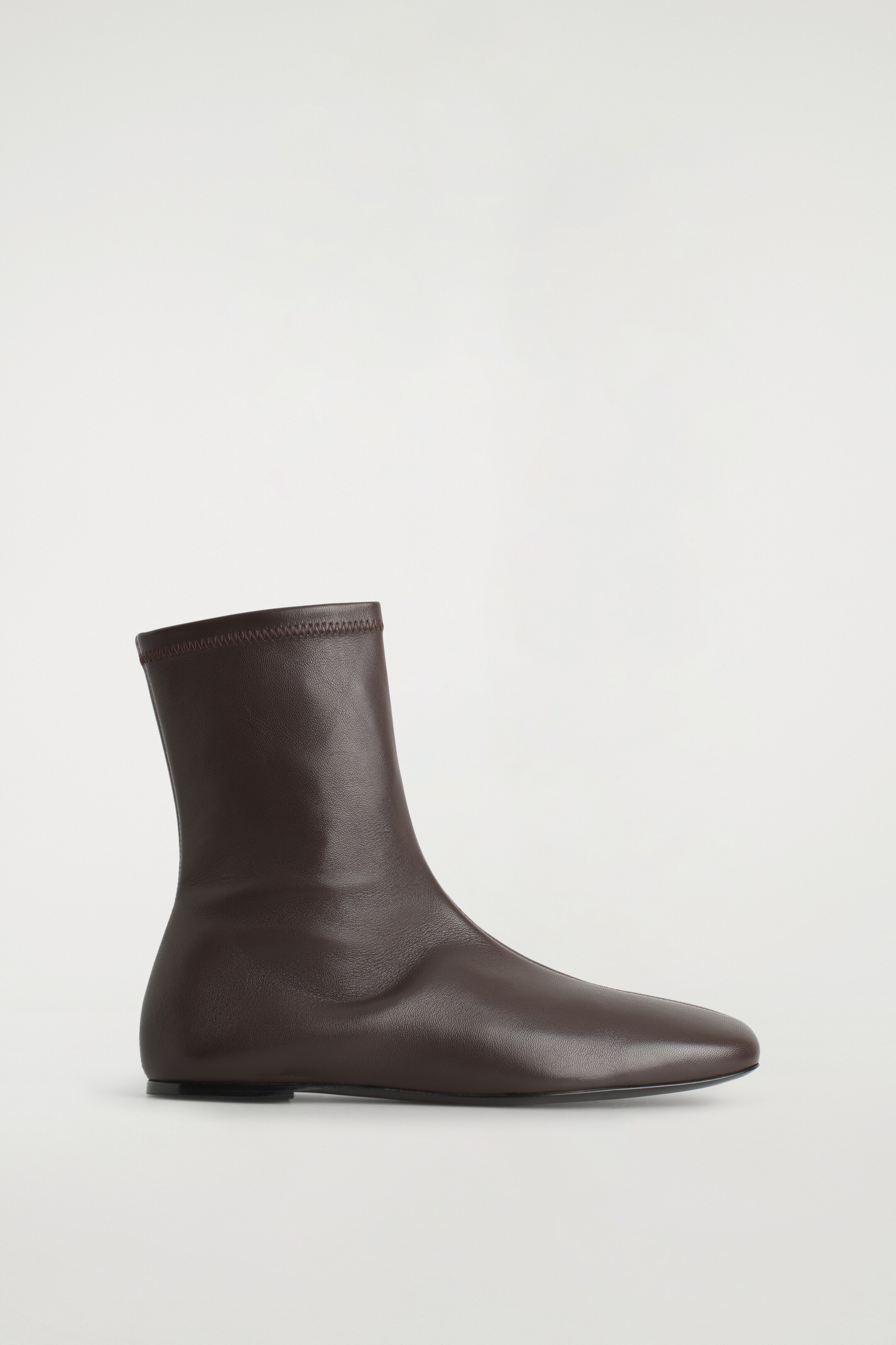 View larger image: A dark brown leather sock boot is standing to the right, featuring a slim, ankle-height shaft with an elasticized opening. It is designed with a rounded square toe and a low heel.