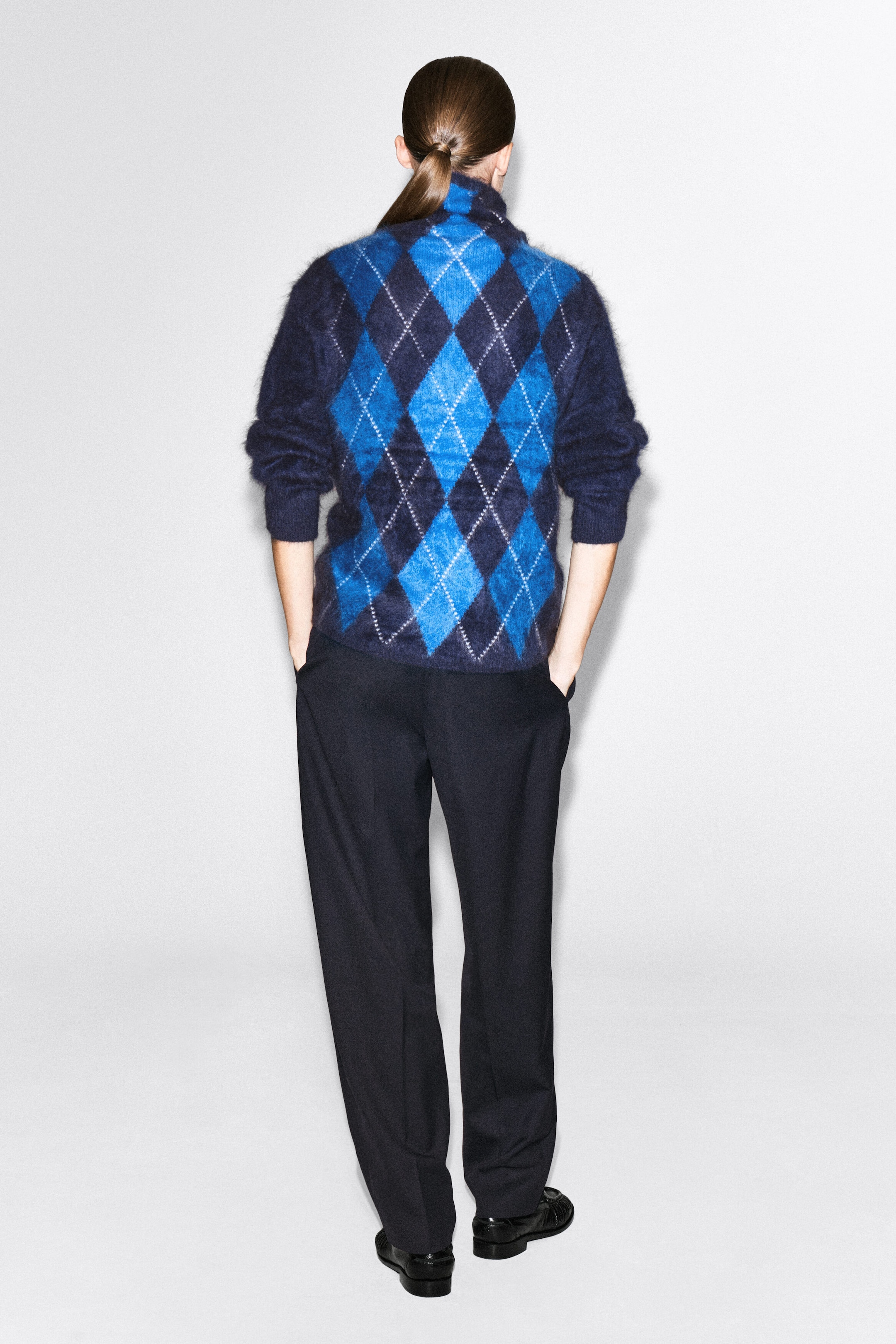 An argyle brushed cashmere funnel-neck jumper in a blue and navy diamond pattern is being worn by the model, who is viewed from the back. The model is styling the jumper with dark trousers, standing with hands resting in the pockets. An argyle brushed cashmere funnel-neck jumper in a blue and navy diamond pattern is being worn by the model, who is viewed from the back. The model is styling the jumper with dark trousers, standing with hands resting in the pockets.