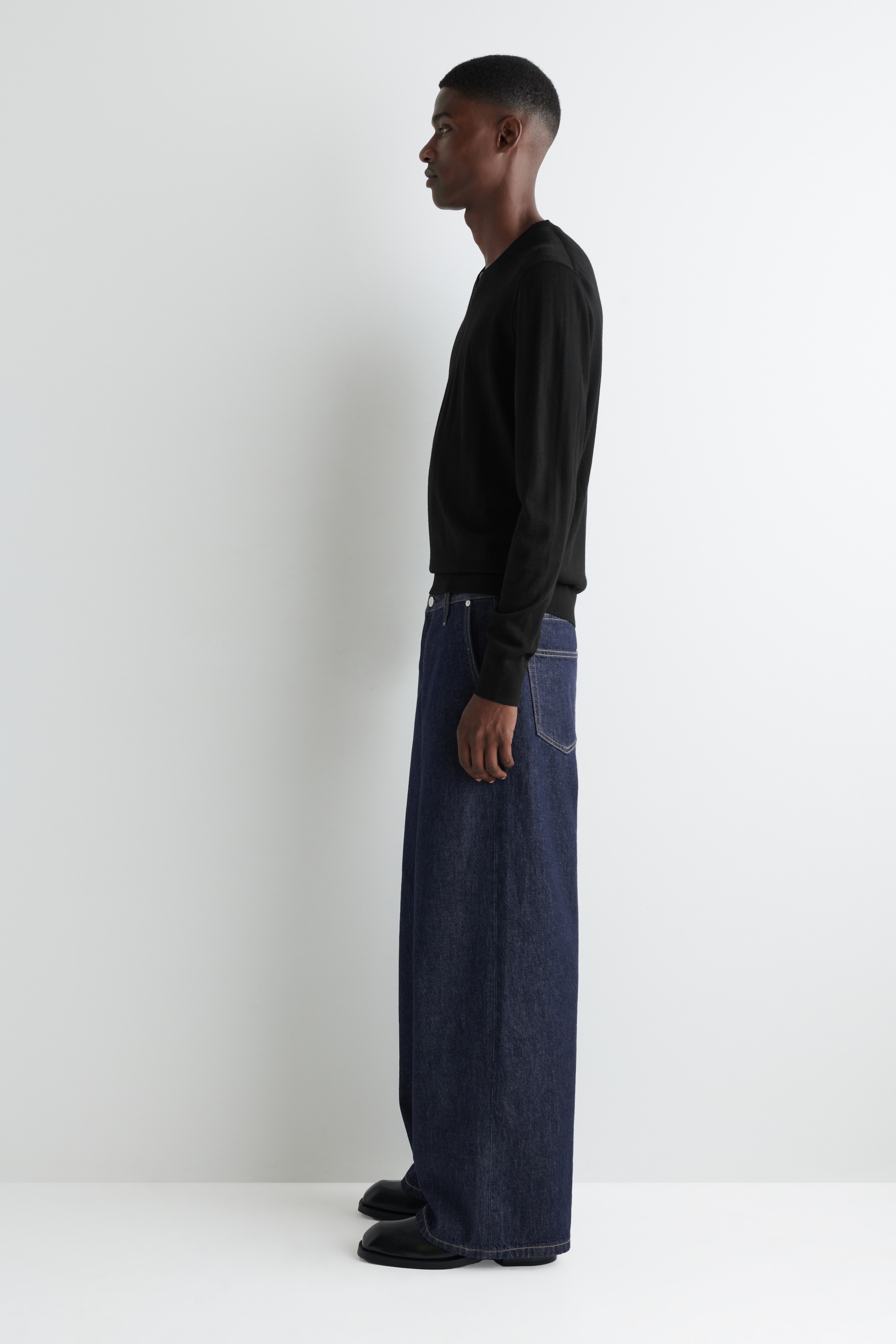 Agrandir l'image: Un haut noir à manches longues et un pantalon en denim bleu foncé à jambes larges sont présentés en vue de profil. Le haut présente un col rond, des poignets côtelés et un ourlet côtelé, tandis que le pantalon en denim à jambes extrêmement larges s'accumule sur des chaussures noires.