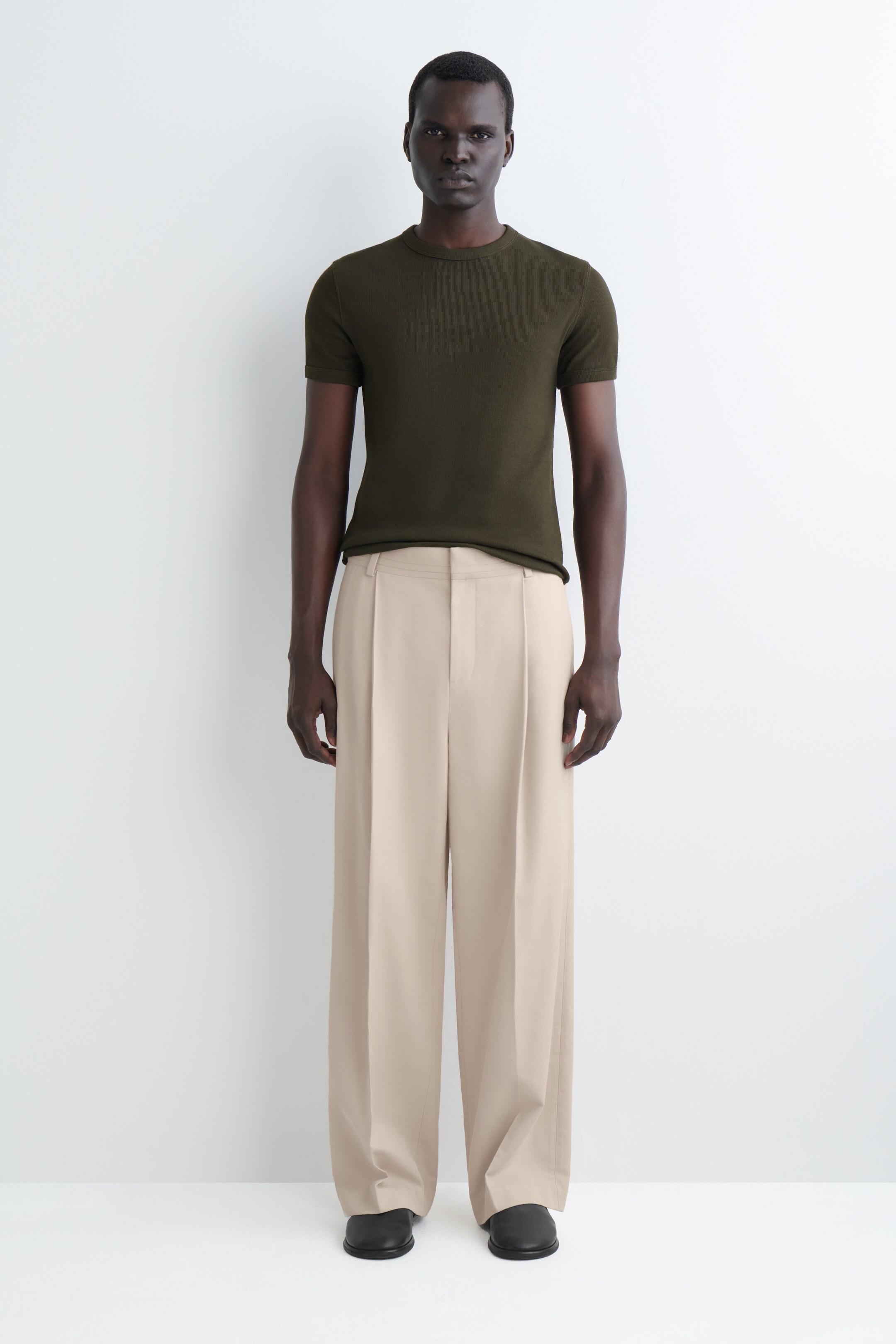 Visualizza immagine più grande: Una maglietta verde oliva scuro, aderente, a maniche corte con collo rotondo viene presentata su una persona che guarda direttamente in avanti. La maglietta è infilata in pantaloni a vita alta, color beige chiaro, a gamba larga, che presentano doppie pieghe sul davanti e si adagiano leggermente sulle caviglie, creando una silhouette rilassata.