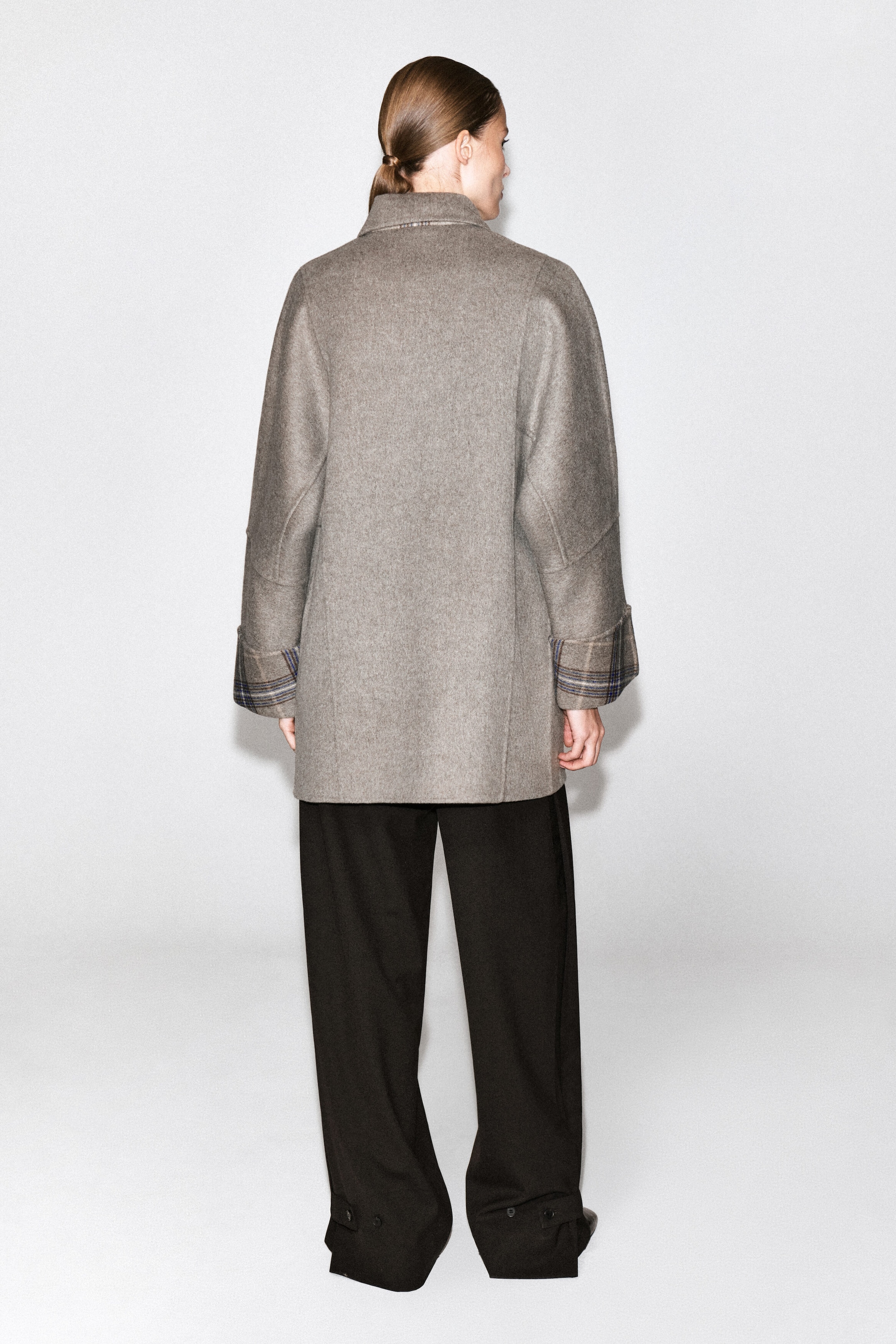 A muted beige wool-blend coat is being worn by the model, shown from the back, styled with loose black trousers. The mid-length coat features a relaxed fit, a high collar, and folded cuffs that reveal a contrasting check pattern.