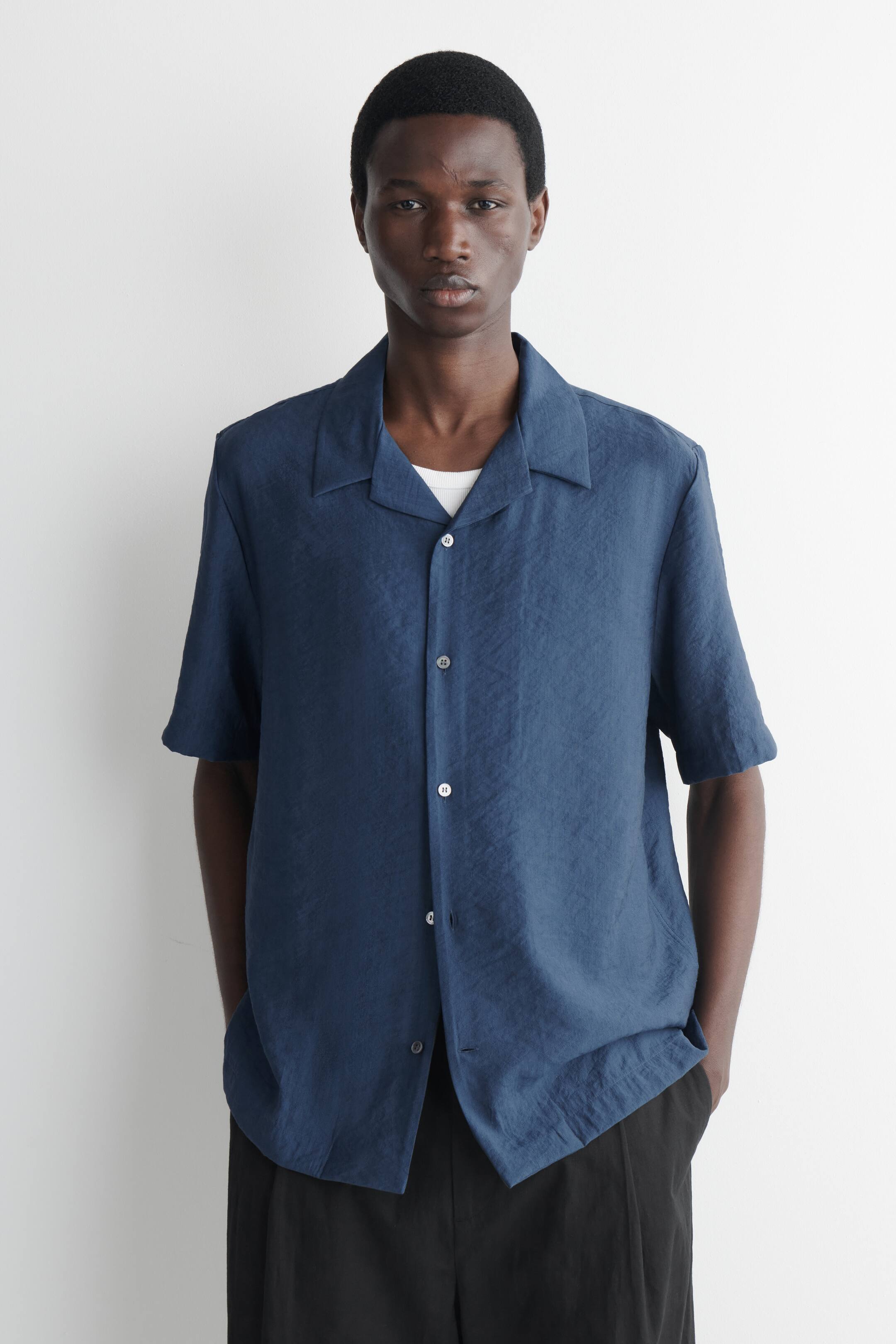 Grösseres Bild ansehen: Ein tiefblaues Kurzarmhemd mit Camp-Kragen und durchgehender Knopfleiste mit weißen Knöpfen. Es zeigt eine subtil geknitterte Textur und wird offen über einem weißen T-Shirt getragen, nach vorne gerichtet.