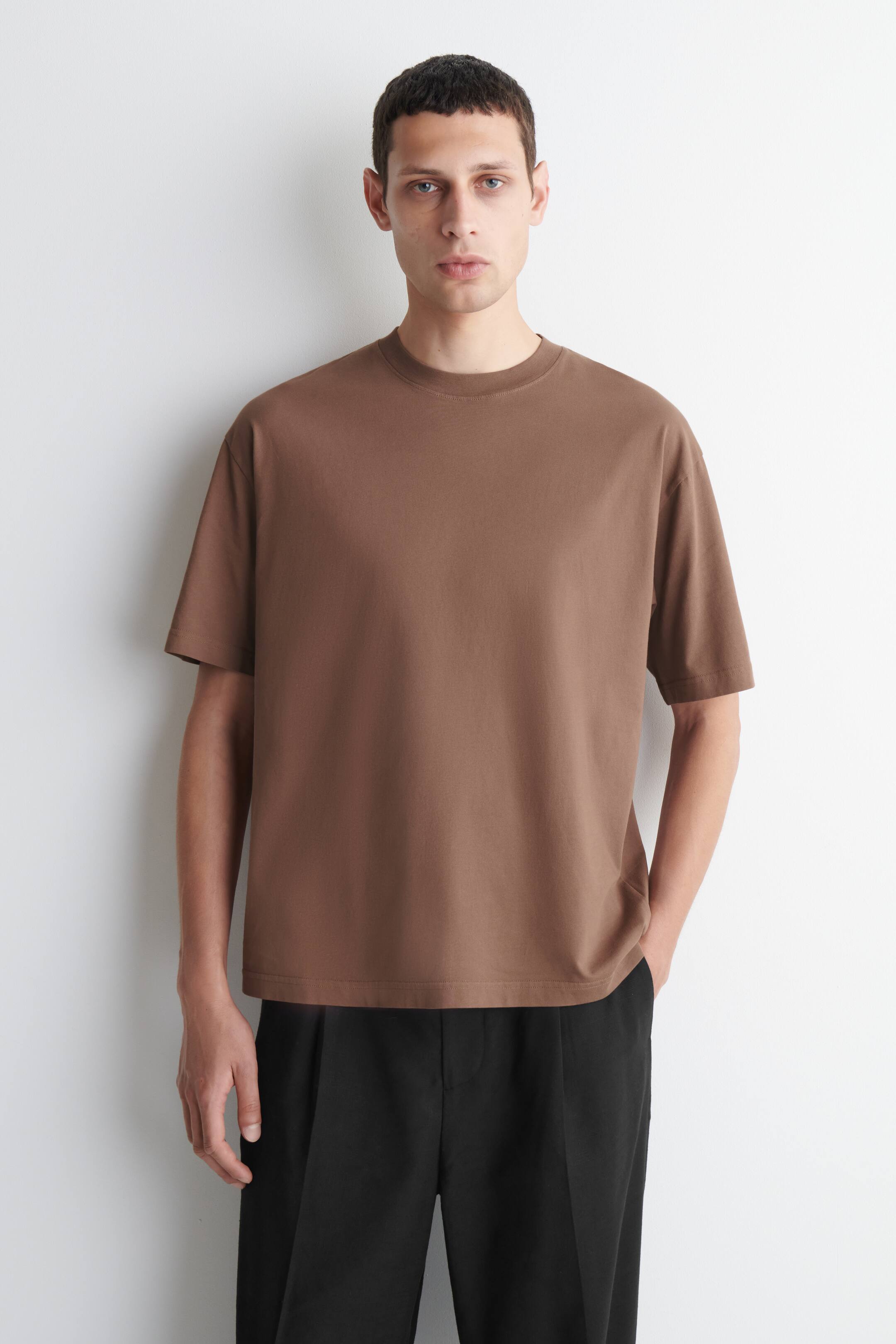 Ver imagem maior: Uma T-shirt castanha de manga curta e gola redonda está a ser usada de frente, exibindo um corte descontraído com ombros caídos.