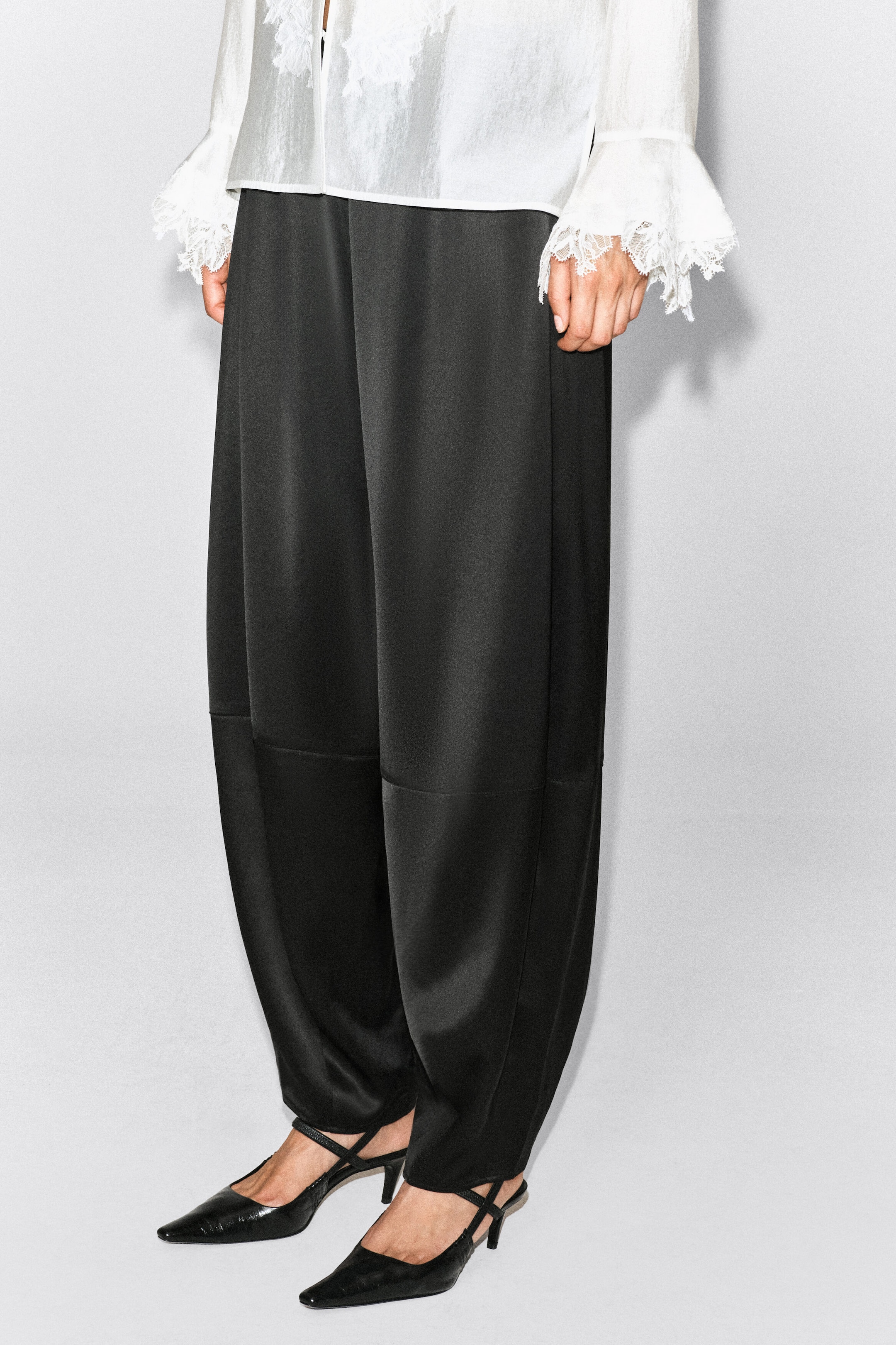 Black satin trousers featuring a barrel-leg silhouette and horizontal seam detailing are worn by the model. The trousers are styled with a white shirt trimmed in lace and black pointed-toe heels. Black satin trousers featuring a barrel-leg silhouette and horizontal seam detailing are worn by the model. The trousers are styled with a white shirt trimmed in lace and black pointed-toe heels.