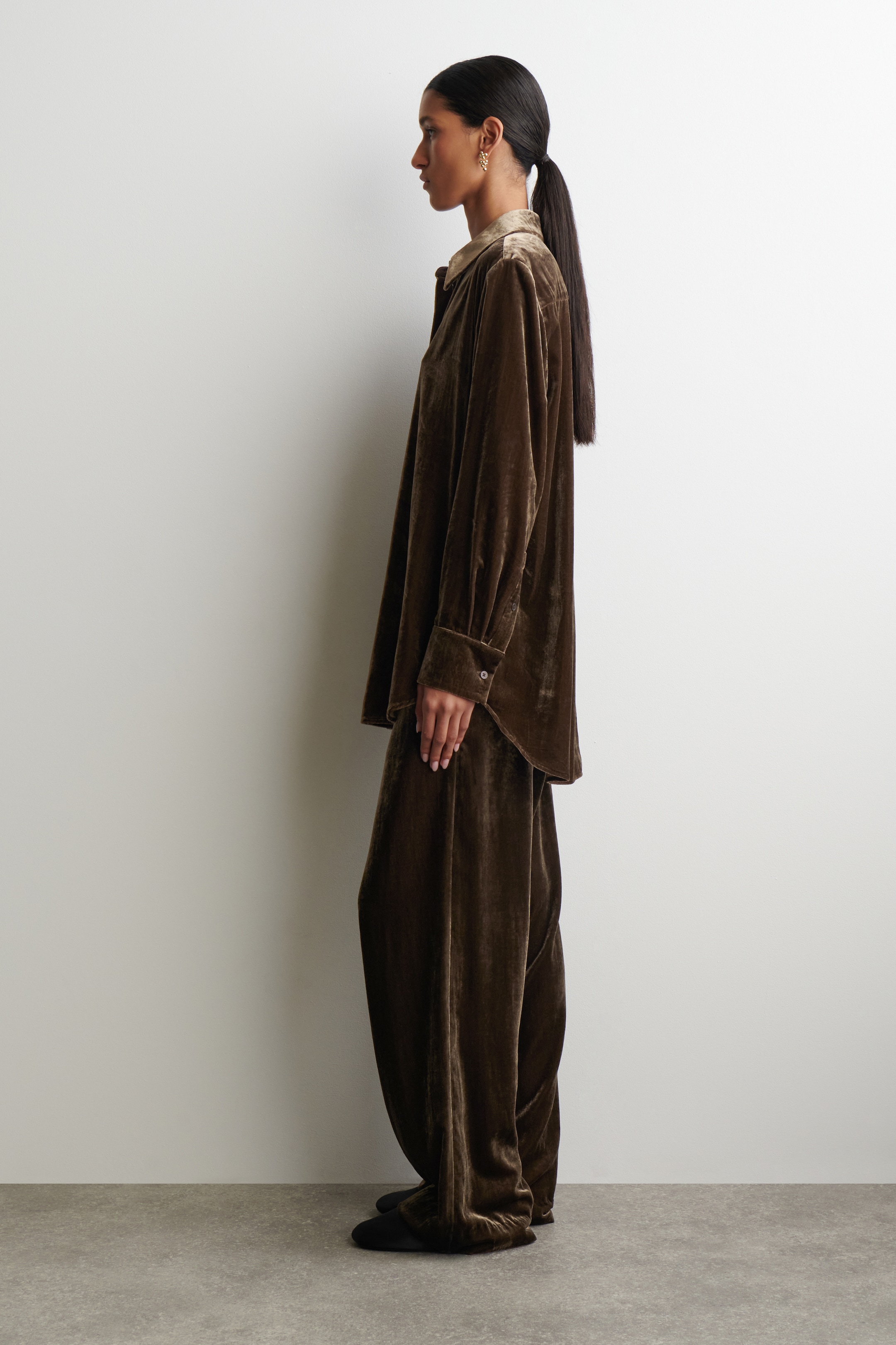 View larger image: A dark brown velvet shirt and matching wide-leg pants are being presented from a side angle. The loose-fitting shirt is featuring a collar and long sleeves with buttoned cuffs, and the full-length pants are falling over dark footwear.