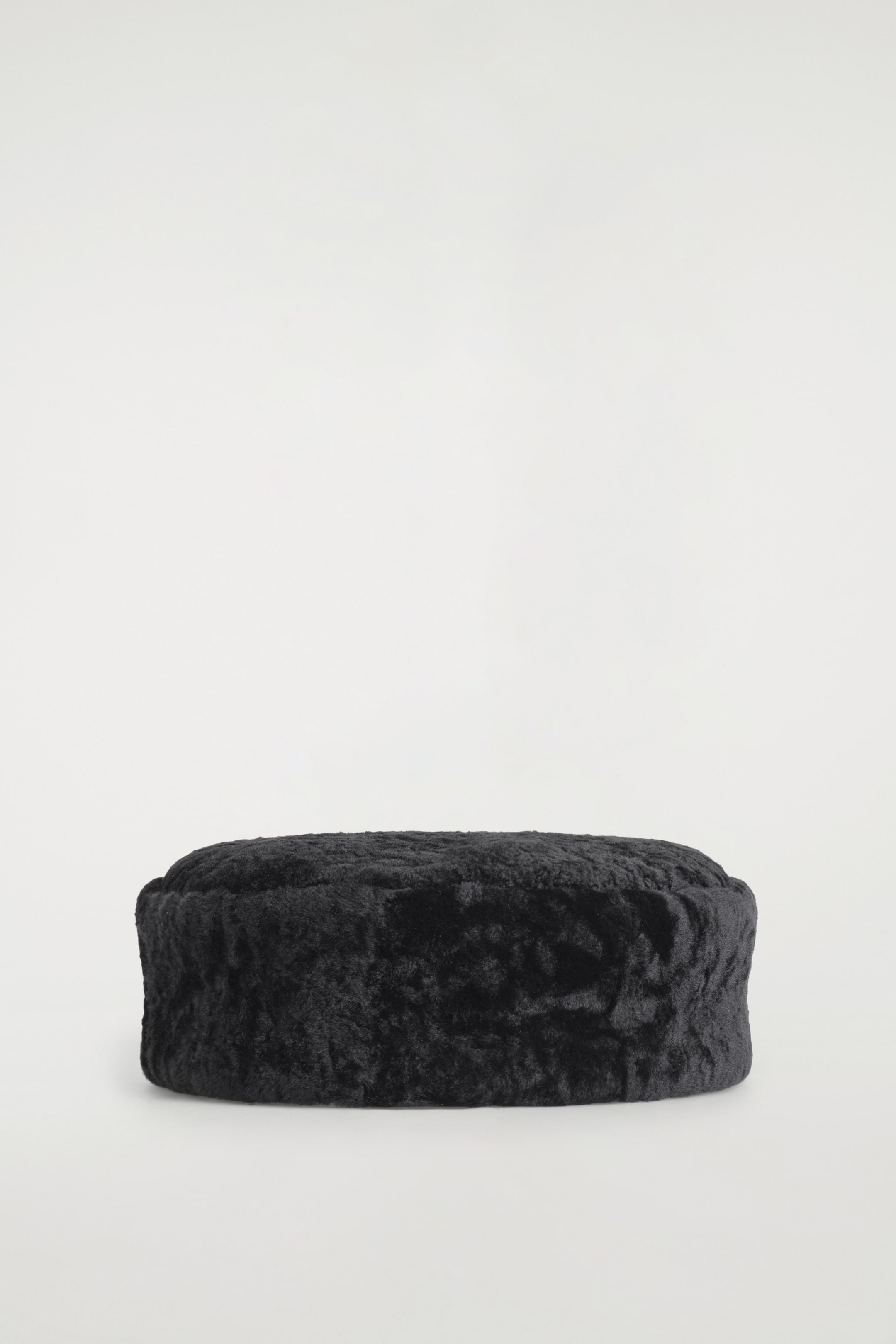 Agrandir l'image: Un chapeau pillbox noir en peau lainée est posé de face, mettant en valeur une surface très texturée et bouclée, avec une forme ronde structurée et un dessus plat.
