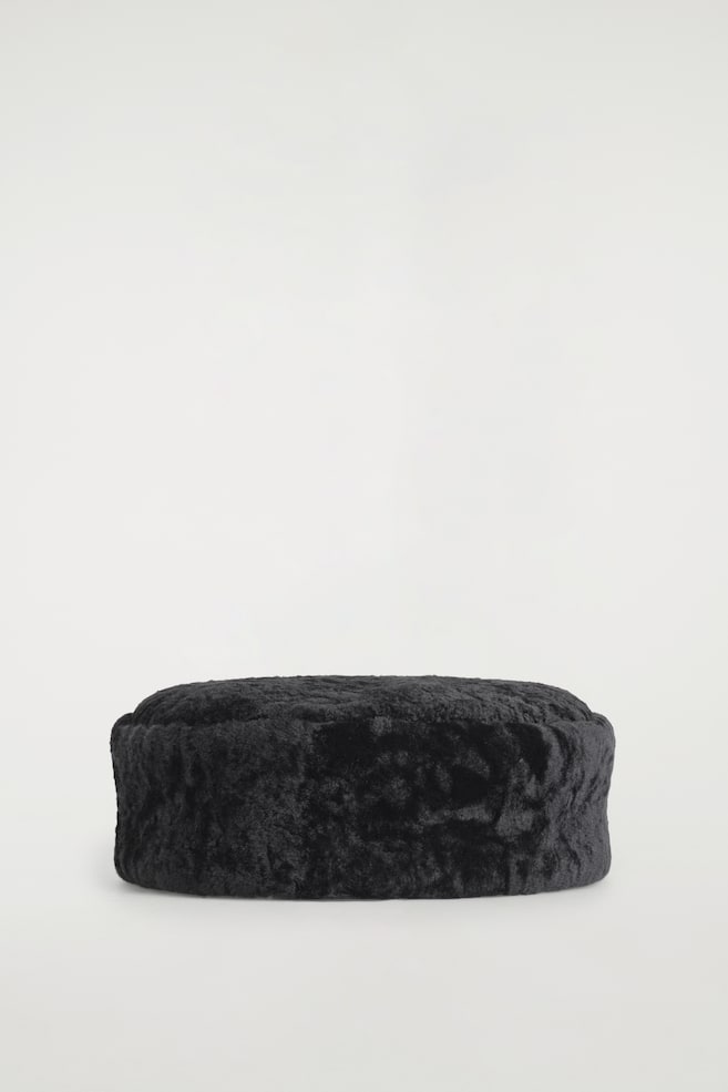 A black shearling pillbox hat is resting facing forwards, showcasing a highly textured, curly surface with a structured, round shape and a flat top. - 1