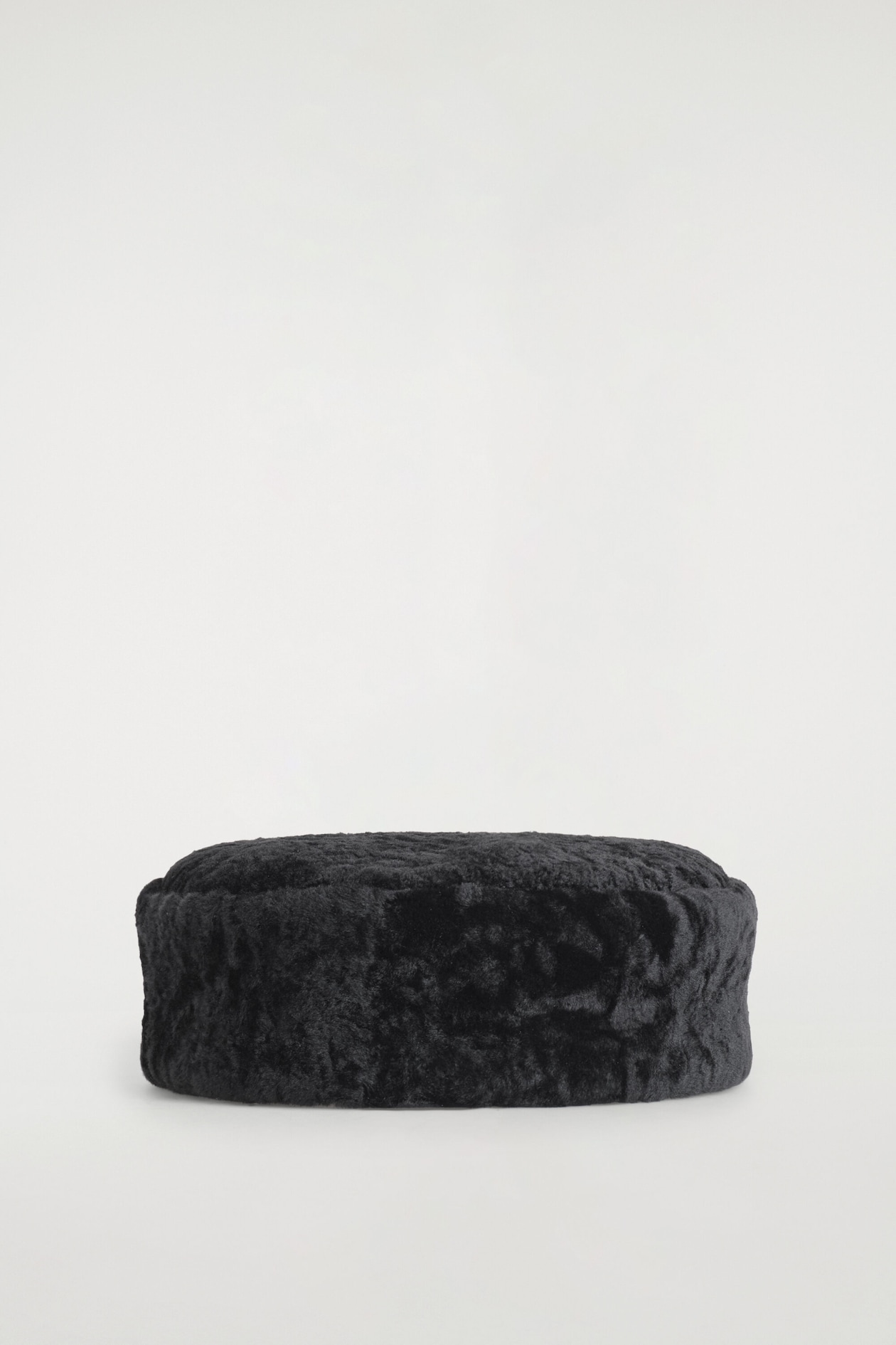A black shearling pillbox hat is resting facing forward, showcasing a highly textured, curly surface with a structured, round shape and a flat top.