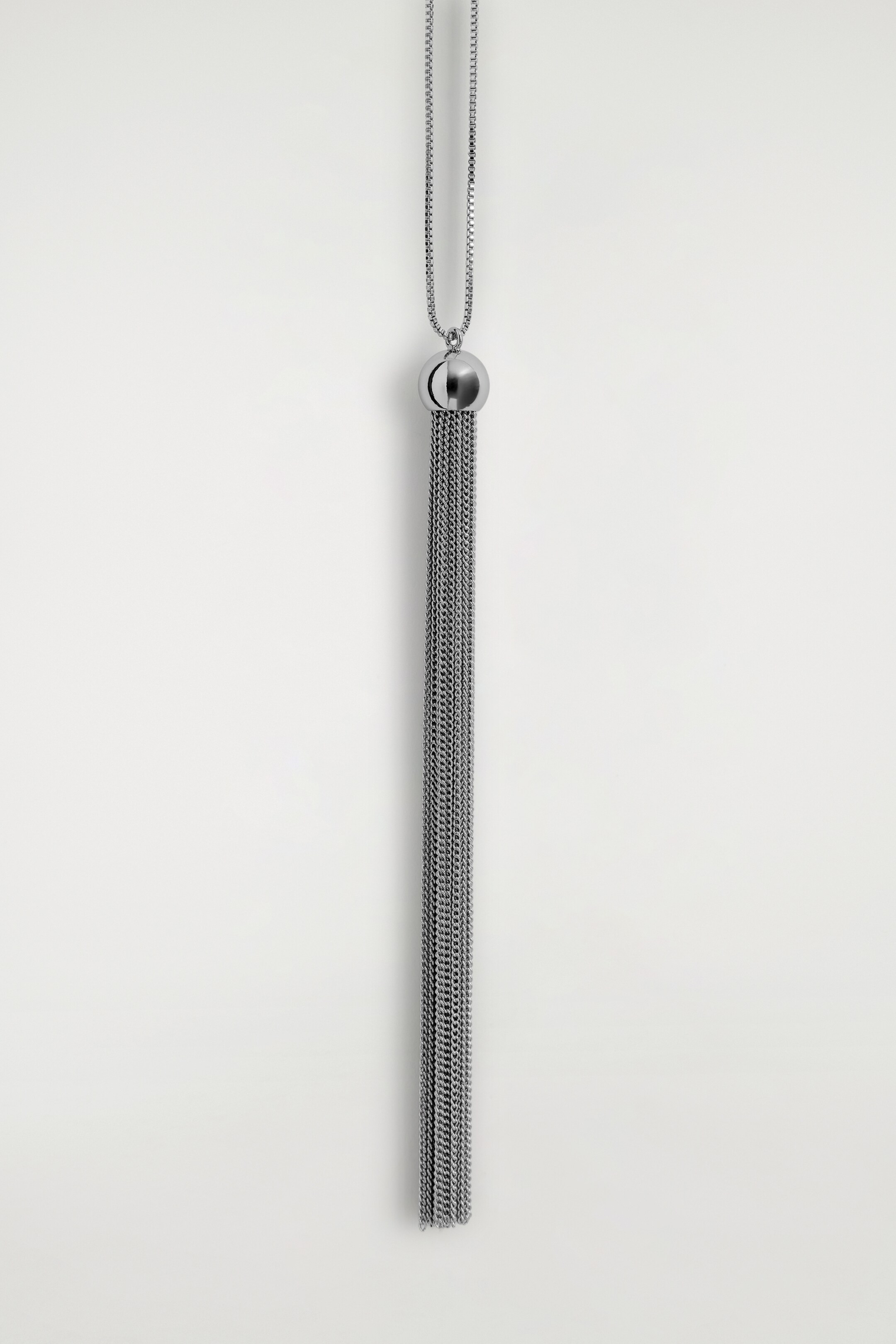 Ver imagen más grande: Un collar de tono plateado está colgando, con una cadena de caja conectada a una cuenta esférica pulida, de la cual múltiples cadenas finas caen en cascada, formando un borla.