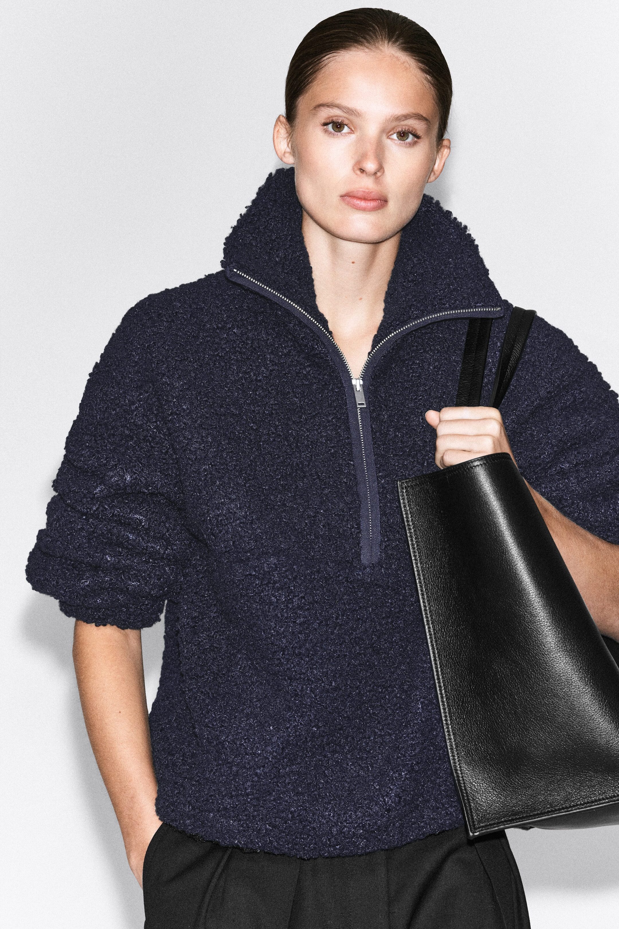 A navy blue half-zip jumper made from textured teddy fabric is worn by the model. The jumper features a high funnel neck and is styled with black trousers and a black leather tote bag carried over the shoulder. A navy blue half-zip jumper made from textured teddy fabric is worn by the model. The jumper features a high funnel neck and is styled with black trousers and a black leather tote bag carried over the shoulder.
