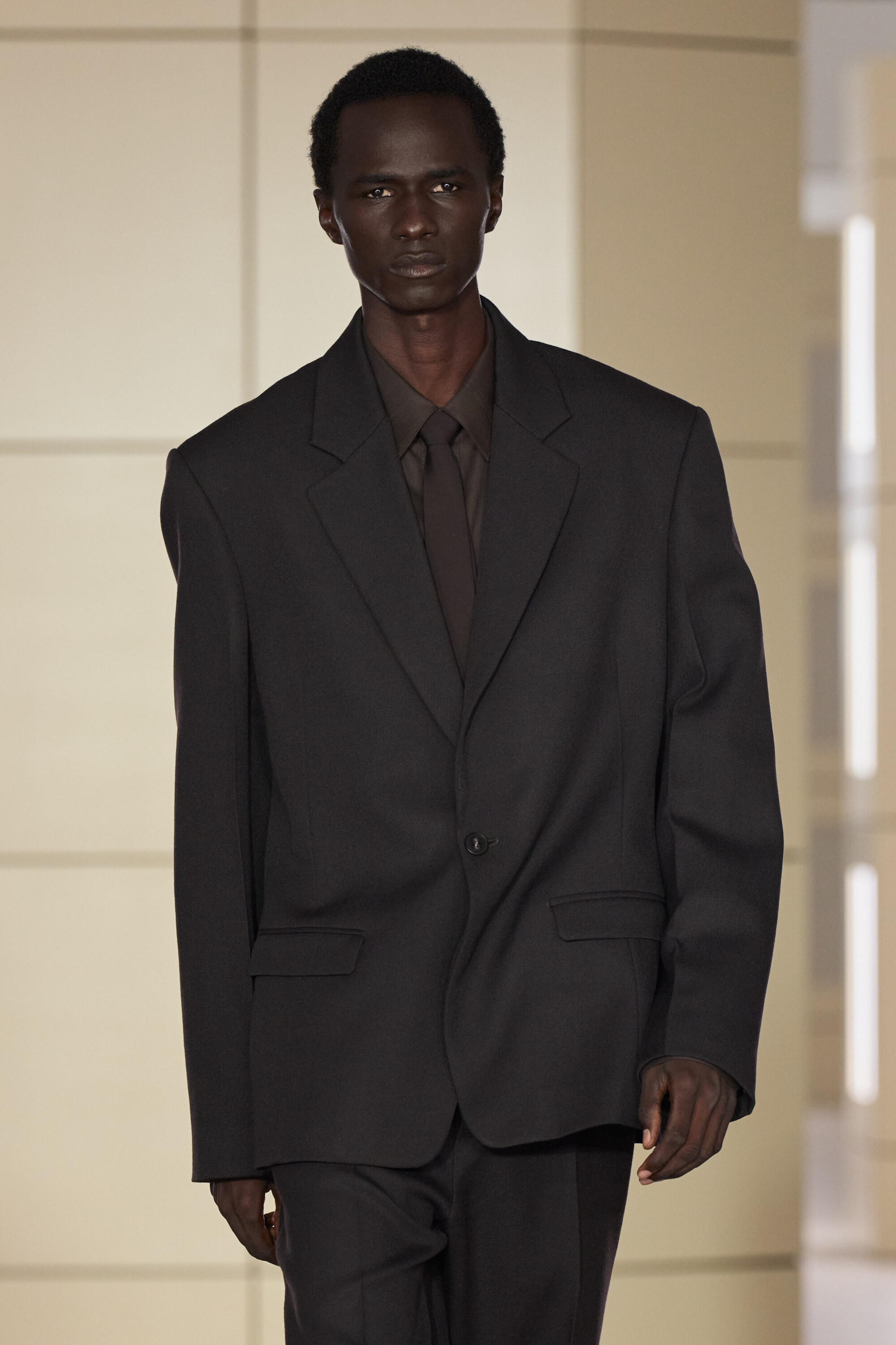 A dark brown wool-twill single-breasted blazer is shown from the front in a structured, oversized fit, featuring notch lapels, padded shoulders, and a single-button fastening. The blazer is layered over a matching collared shirt and tie, highlighting its classic flap pockets. A dark brown wool-twill single-breasted blazer is shown from the front in a structured, oversized fit, featuring notch lapels, padded shoulders, and a single-button fastening. The blazer is layered over a matching collared shirt and tie, highlighting its classic flap pockets.