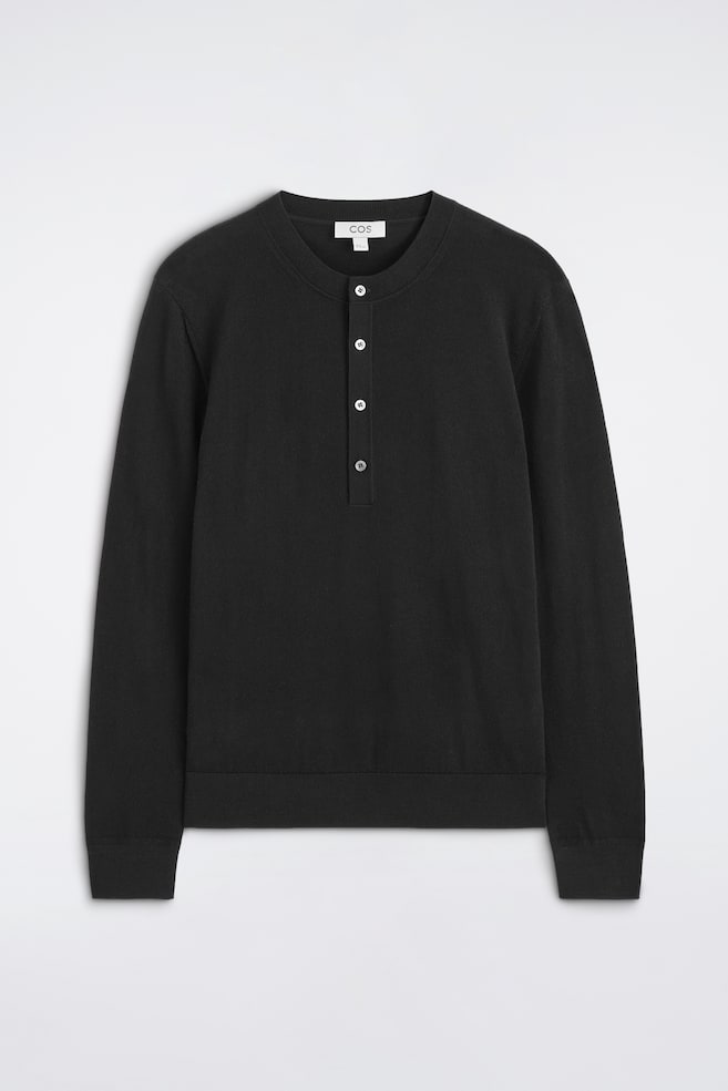 Se muestra de frente un top Henley negro de manga larga. Presenta una textura de punto fino, un escote redondo con tapeta de botones y cinco botones de color claro. El top está rematado con puños acanalados y un bajo acanalado. - 1
