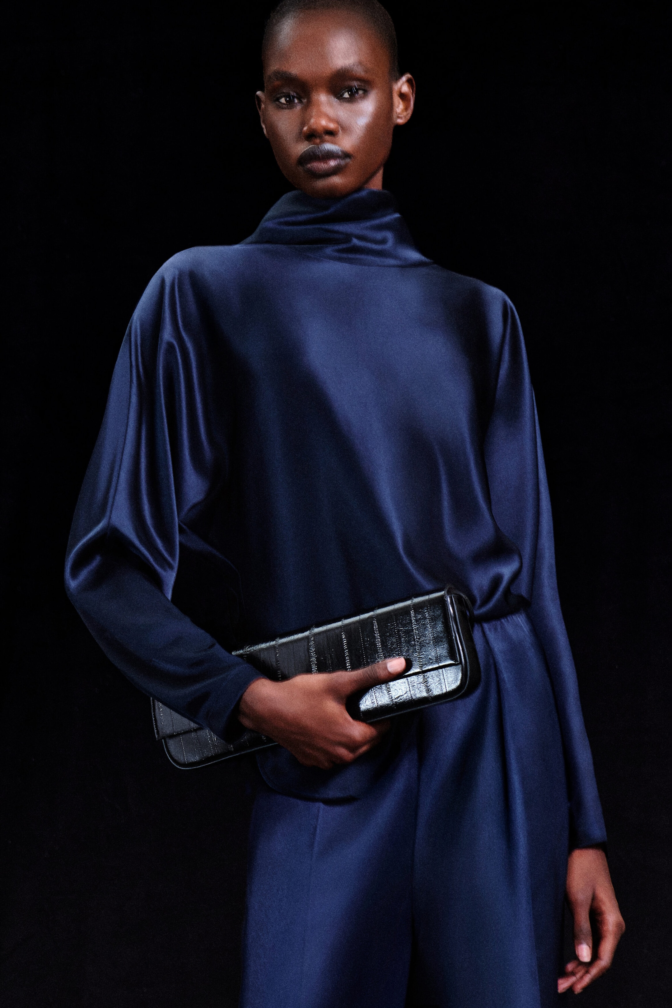 The navy blue cowl-neck satin blouse and matching satin trousers drape softly, creating a fluid silhouette from the front. The long-sleeved blouse features a high, draped cowl neckline, and the model is holding a black textured clutch bag across the waist. The navy blue cowl-neck satin blouse and matching satin trousers drape softly, creating a fluid silhouette from the front. The long-sleeved blouse features a high, draped cowl neckline, and the model is holding a black textured clutch bag across the waist.