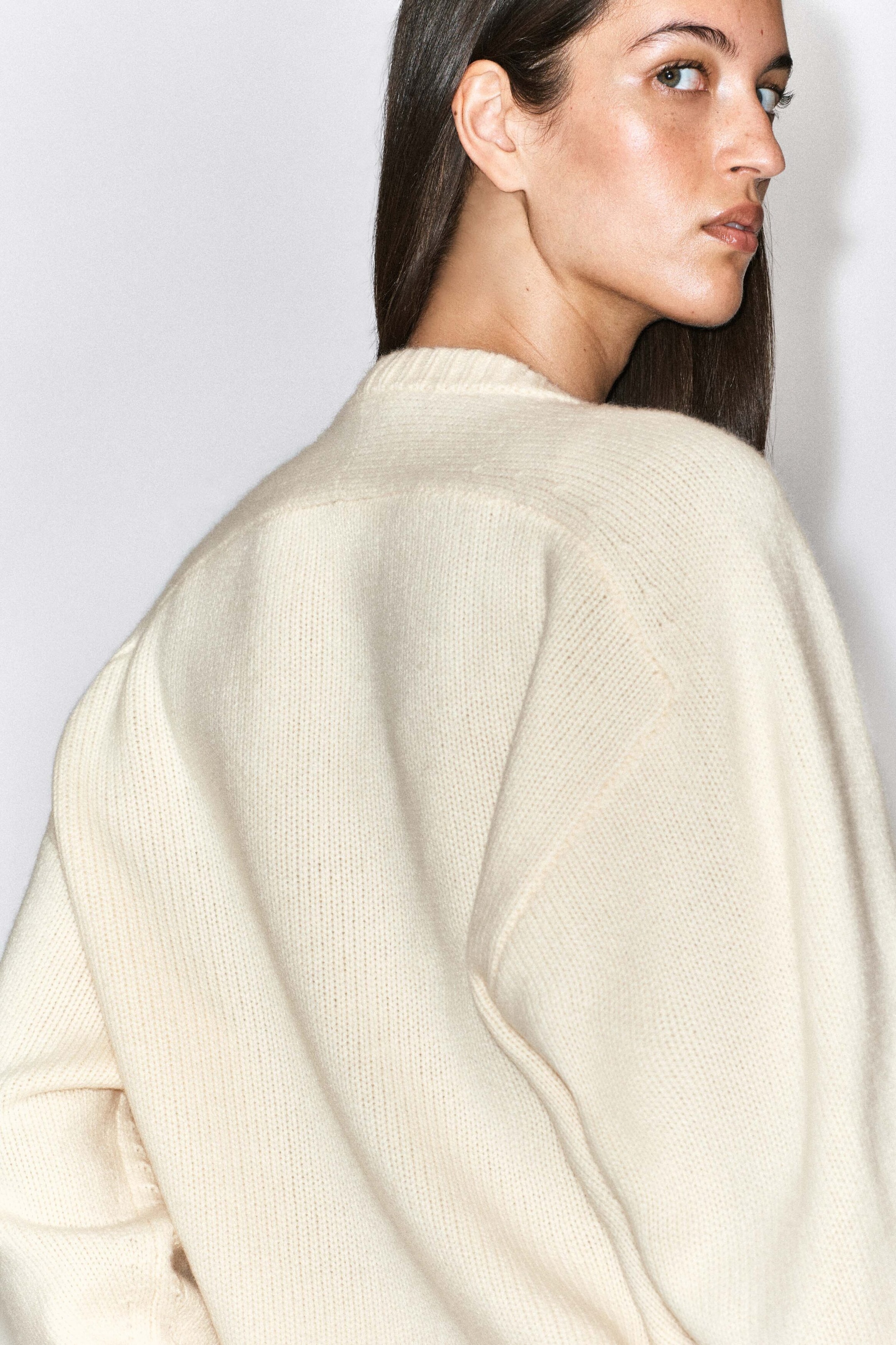 A cream-coloured cashmere crew-neck jumper with a soft knit texture is being worn by the model, showing the rounded shoulder construction and ribbed collar from behind. A cream-coloured cashmere crew-neck jumper with a soft knit texture is being worn by the model, showing the rounded shoulder construction and ribbed collar from behind.