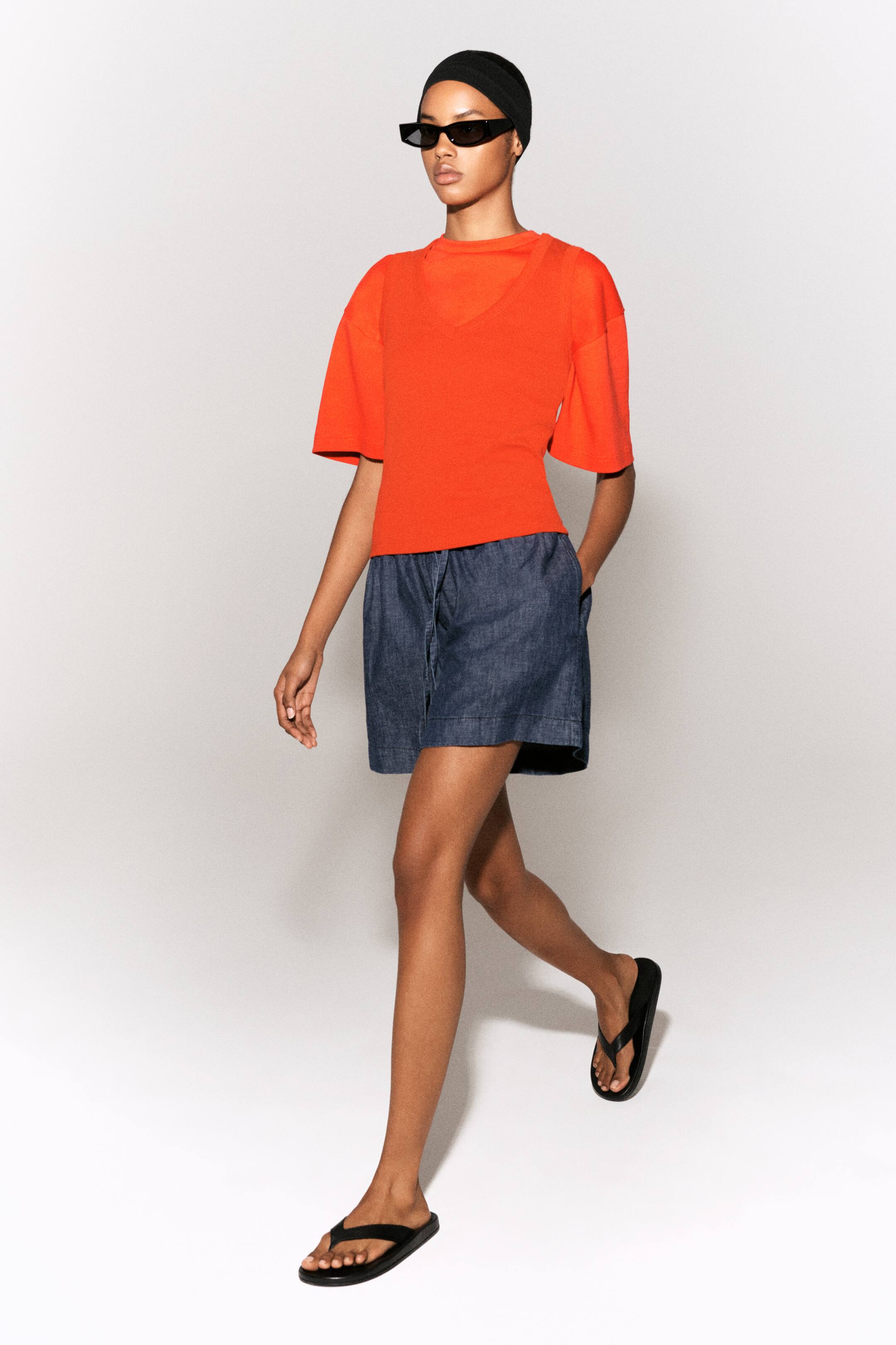 Agrandir l'image: Shorts bleu marine avec une taille élastiquée à cordon et des poches latérales portés avec un haut à manches courtes de forme carrée, de couleur orange vif, avec des épaules tombantes, porté par-dessus un débardeur à col en V couleur mandarine.