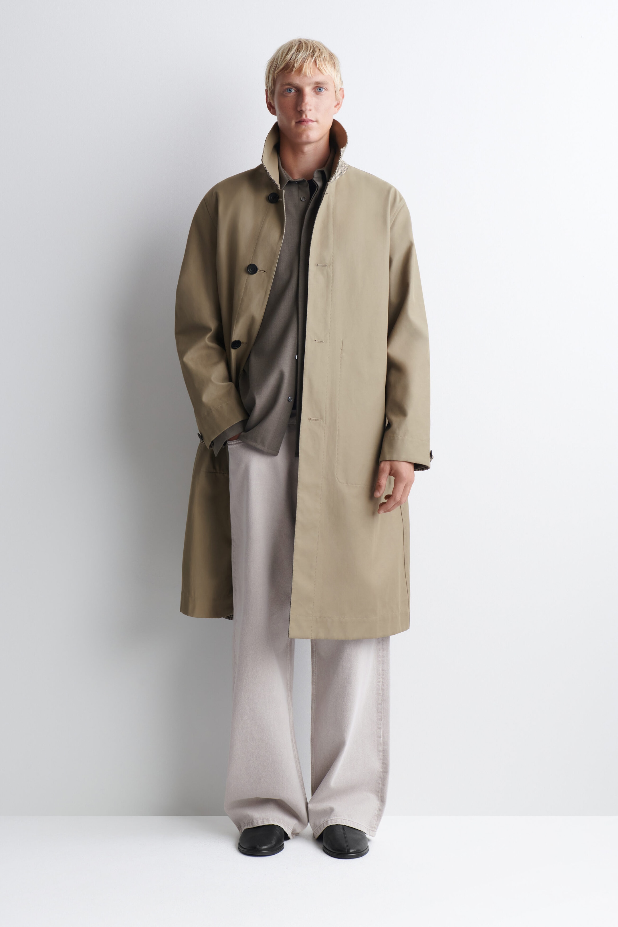 Le manteau de voiture réversible à carreaux est présenté de face, mettant en valeur son extérieur beige, sa silhouette décontractée jusqu’aux genoux et son col montant qui laisse apparaître un motif à carreaux sur l’envers. Il est porté ouvert sur une chemise grise boutonnée et un pantalon clair, soulignant sa fermeture à simple boutonnage et son ourlet droit. Le manteau de voiture réversible à carreaux est présenté de face, mettant en valeur son extérieur beige, sa silhouette décontractée jusqu’aux genoux et son col montant qui laisse apparaître un motif à carreaux sur l’envers. Il est porté ouvert sur une chemise grise boutonnée et un pantalon clair, soulignant sa fermeture à simple boutonnage et son ourlet droit.