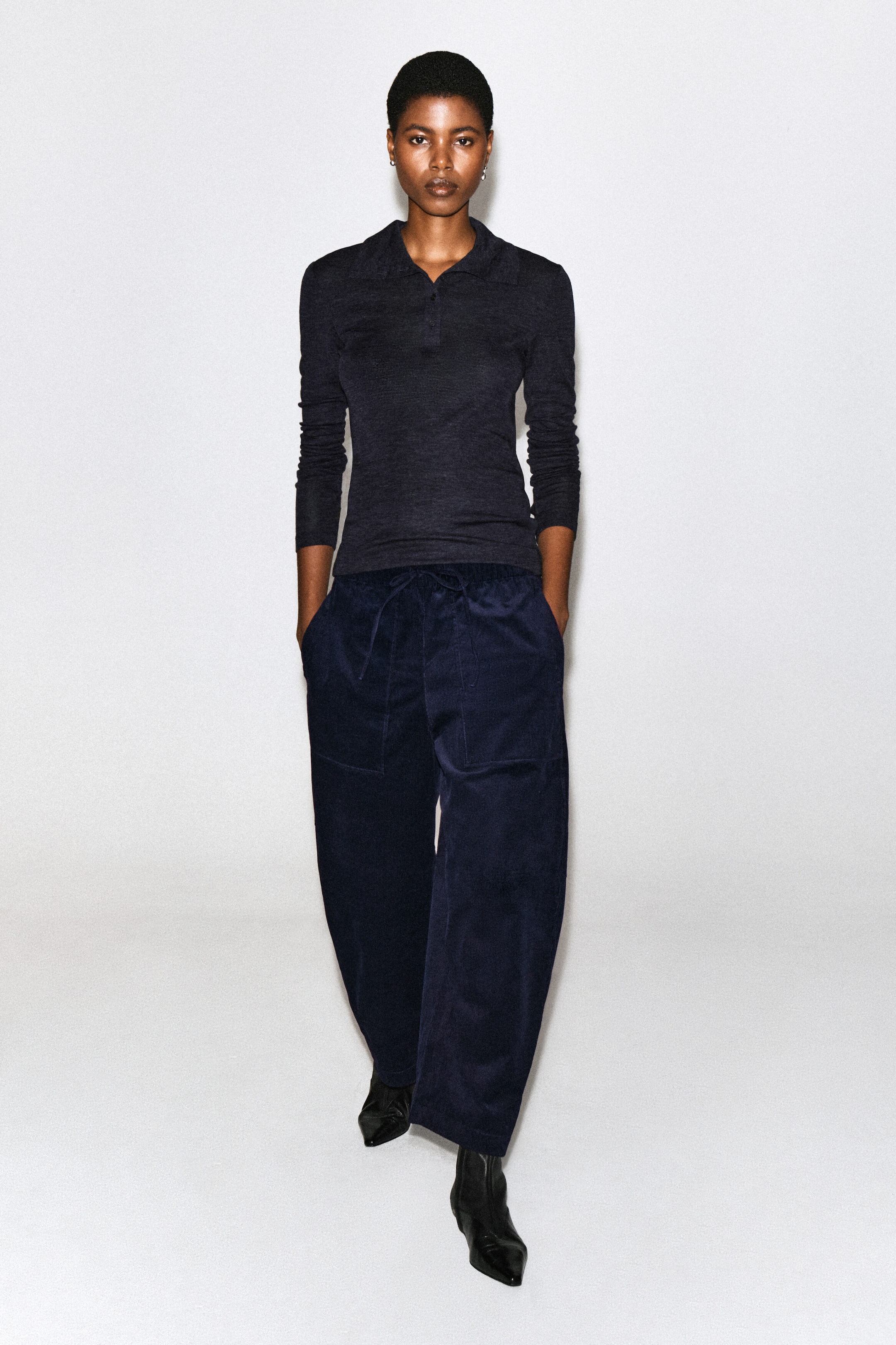 Corduroy barrel-leg drawstring pants in dark navy blue are worn by the model, who is posing with her hands in the front pockets. The pants feature a relaxed, curved-leg silhouette and a drawstring waist, and are styled with a dark long-sleeve polo shirt and black pointed-toe boots. Corduroy barrel-leg drawstring pants in dark navy blue are worn by the model, who is posing with her hands in the front pockets. The pants feature a relaxed, curved-leg silhouette and a drawstring waist, and are styled with a dark long-sleeve polo shirt and black pointed-toe boots.