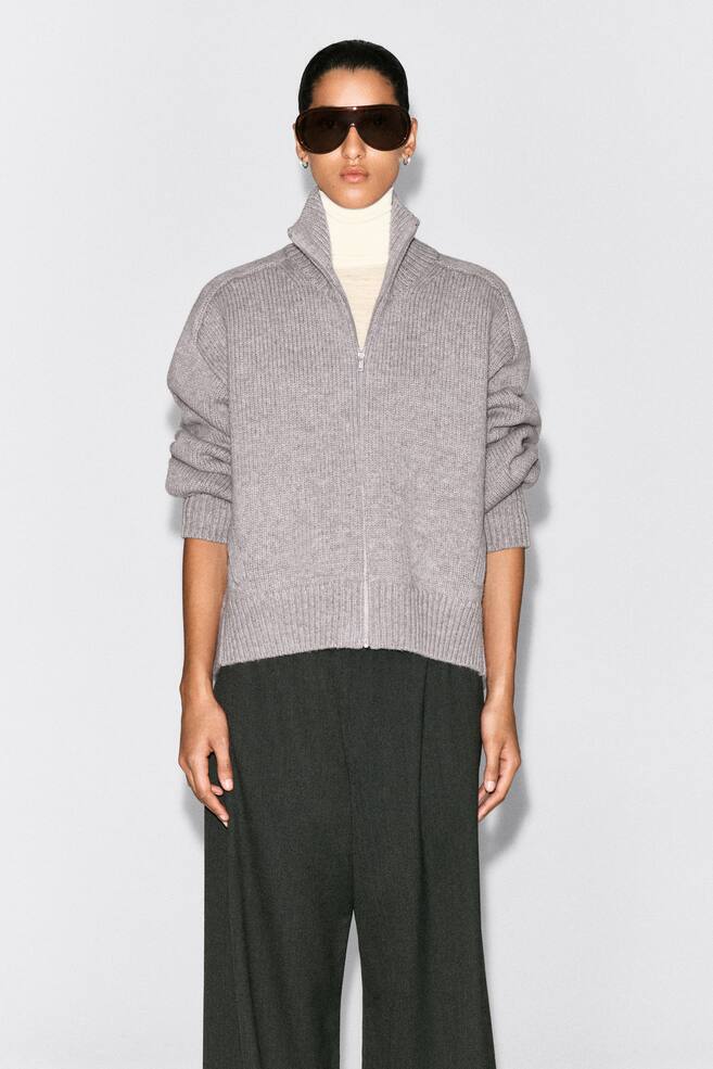 Die Jacke mit Rundhalsausschnitt aus Wolle und ReiÃverschluss wird von vorne gezeigt und zeichnet sich durch eine grau gerippte Strickstruktur, einen silbernen FrontreiÃverschluss und einen hohen Kragen aus. Sie wird über einem cremefarbenen Rollkragenpullover getragen und betont ihre lockere Passform, die überschnittenen Schultern sowie gerippte Bündchen und Saum. - 1