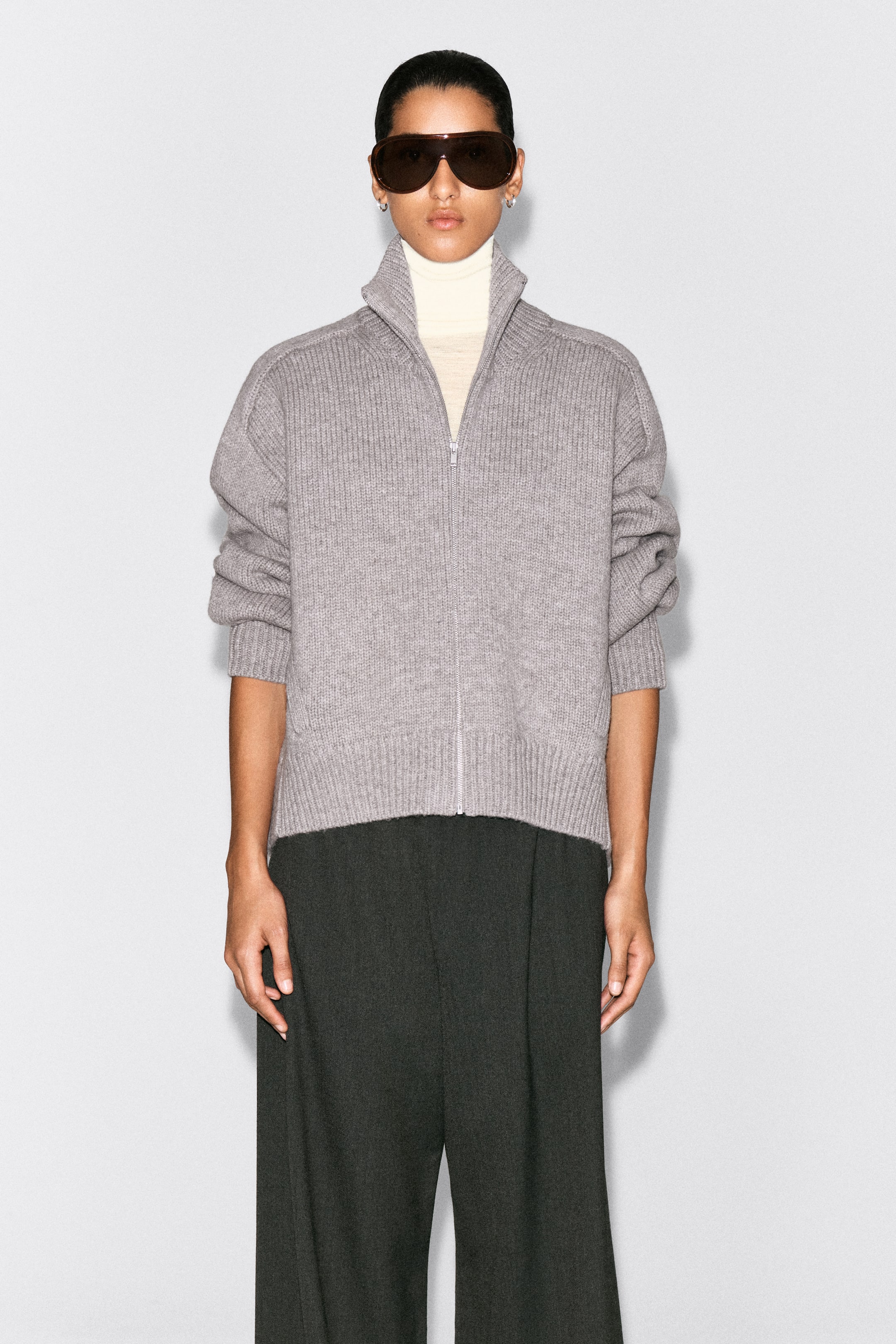 Visualizza immagine più grande: La giacca zip-up in lana con collo a imbuto arrotondato è mostrata frontalmente, con una texture a maglia a coste grigio, una cerniera frontale argentata e un colletto alto. È indossata sopra un dolcevita color crema, evidenziando la vestibilità rilassata, le spalle scese e i polsini e l’orlo a costine.