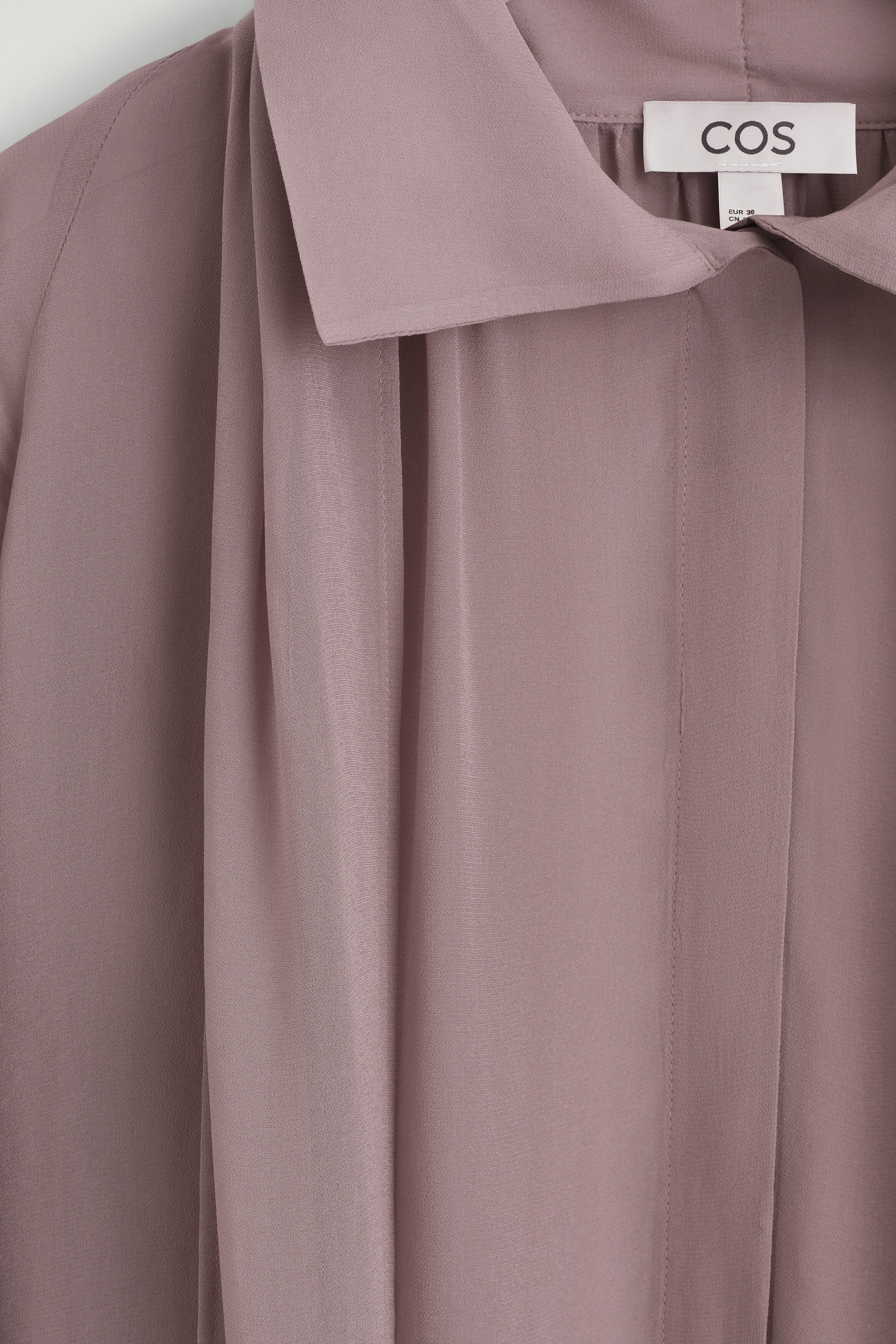 Un gros plan du haut fluide à détail foulard dans une teinte mauve poussiéreuse met en valeur son col pointu ainsi qu’un panneau de tissu drapé qui cascade sur le devant. Le haut présente une texture légère et fluide, avec une patte de boutonnage dissimulée sur le devant et une étiquette de marque visible à l’intérieur du col.