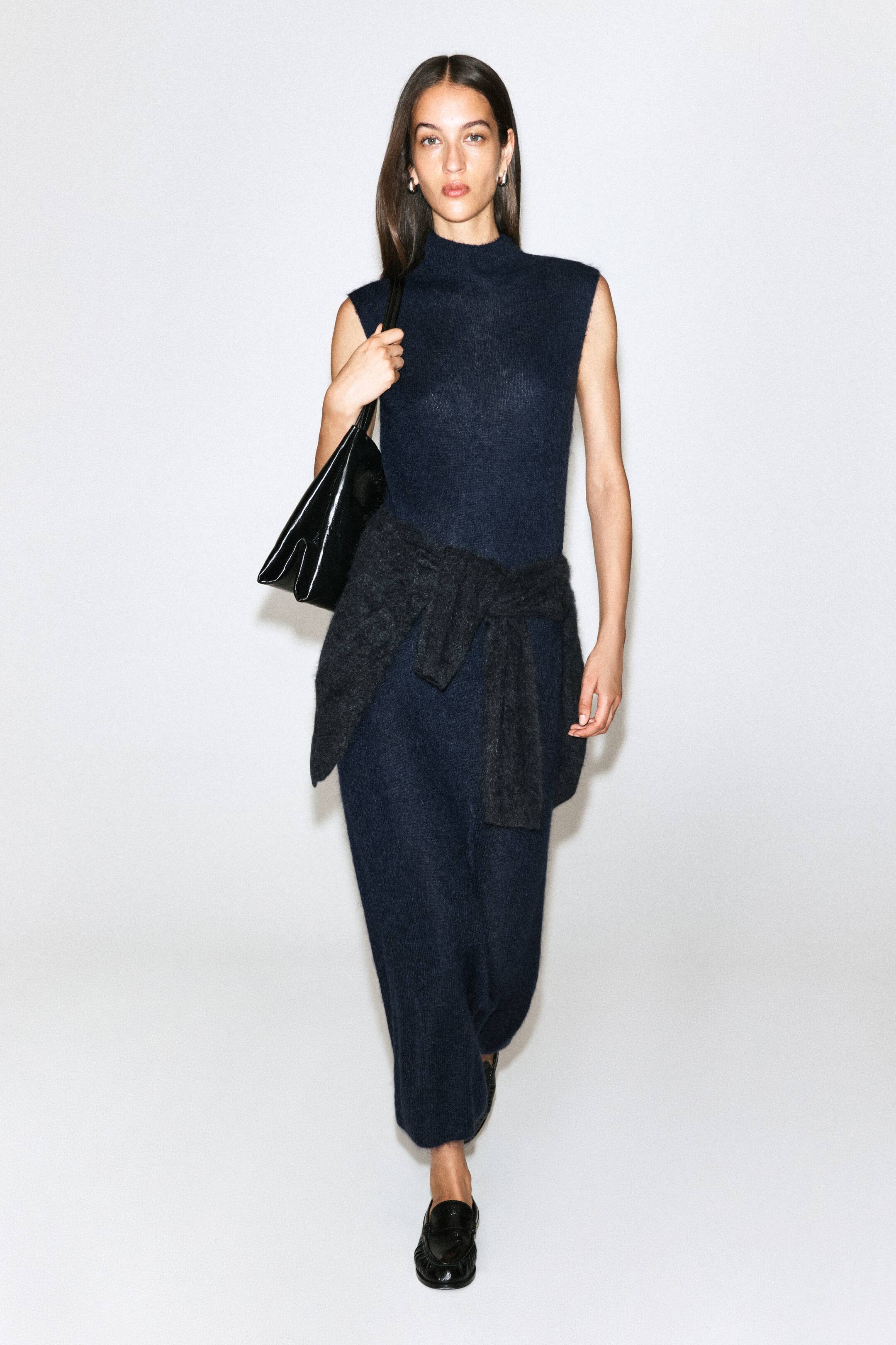 Ver imagen más grande: La modelo lleva un vestido de cuello chimenea cepillado en azul marino oscuro, con silueta midi recta y sin mangas. El vestido de punto se combina con un jersey gris oscuro anudado a la cintura y se acompaña de un bolso negro de hombro y unos mocasines negros.