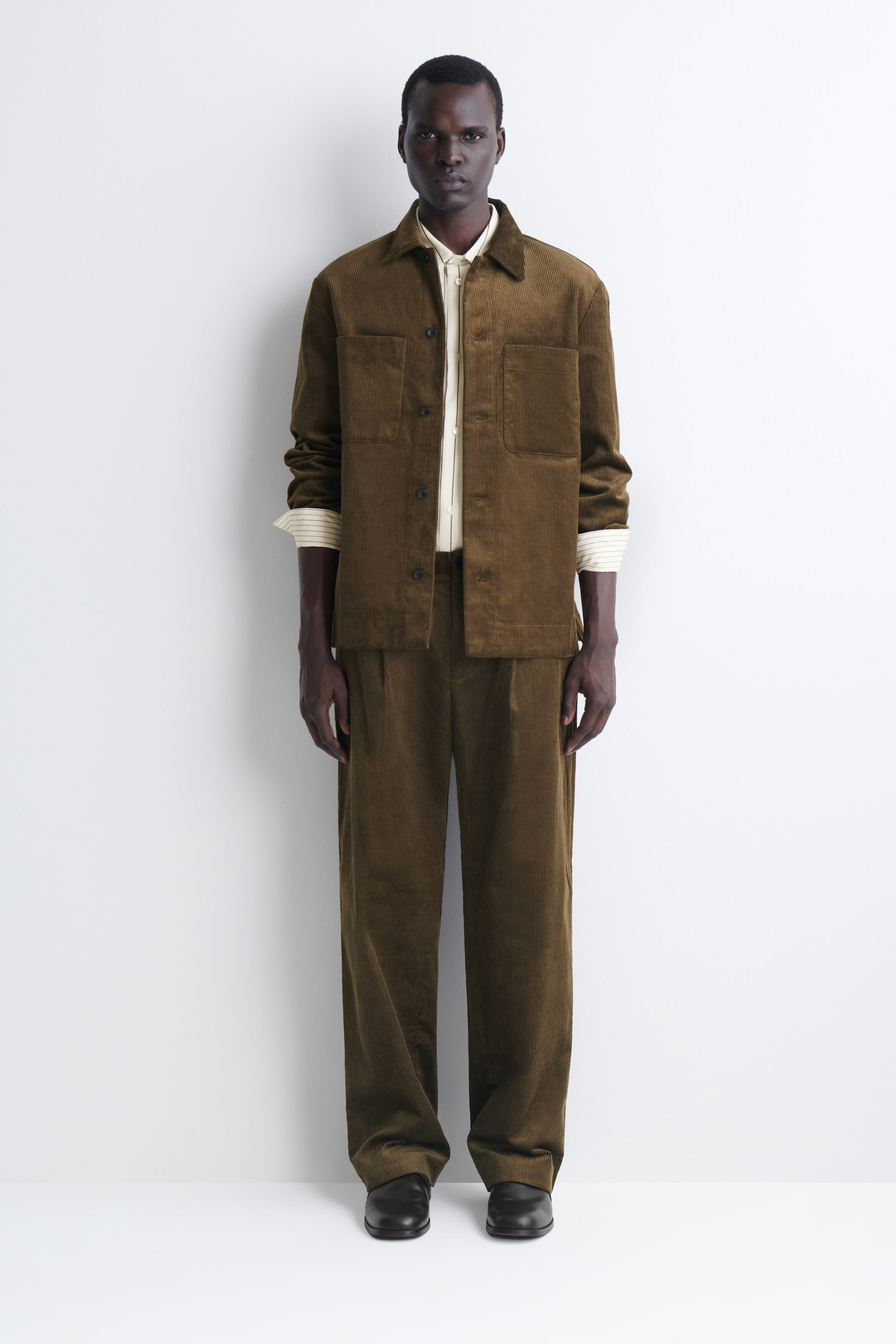 The Pleated Relaxed-Fit Tapered Corduroy Pants in a dark olive-brown color are shown from the front, featuring a relaxed silhouette with subtle front pleats and a tapered leg. The pants drape over black leather shoes and are styled with a matching corduroy jacket to highlight their textured finish. The Pleated Relaxed-Fit Tapered Corduroy Pants in a dark olive-brown color are shown from the front, featuring a relaxed silhouette with subtle front pleats and a tapered leg. The pants drape over black leather shoes and are styled with a matching corduroy jacket to highlight their textured finish.