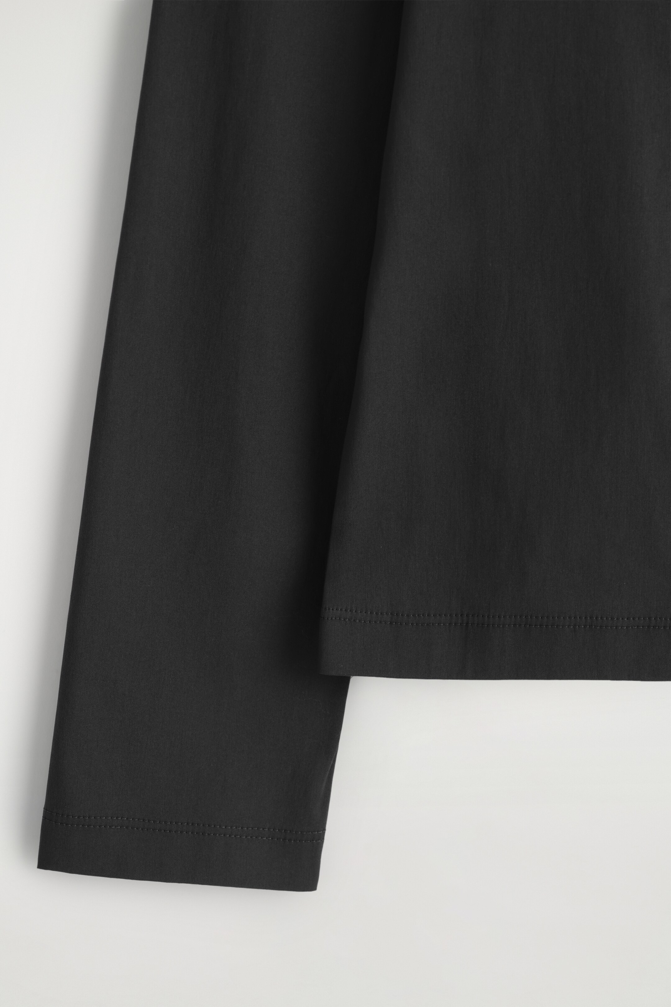 View larger image: A black long-sleeved top is being shown in a close-up, highlighting its smooth fabric and refined construction. The front of the top is featuring a straight bottom hem, and a long sleeve is ending in a simple cuff, both with visible double-stitched detailing.