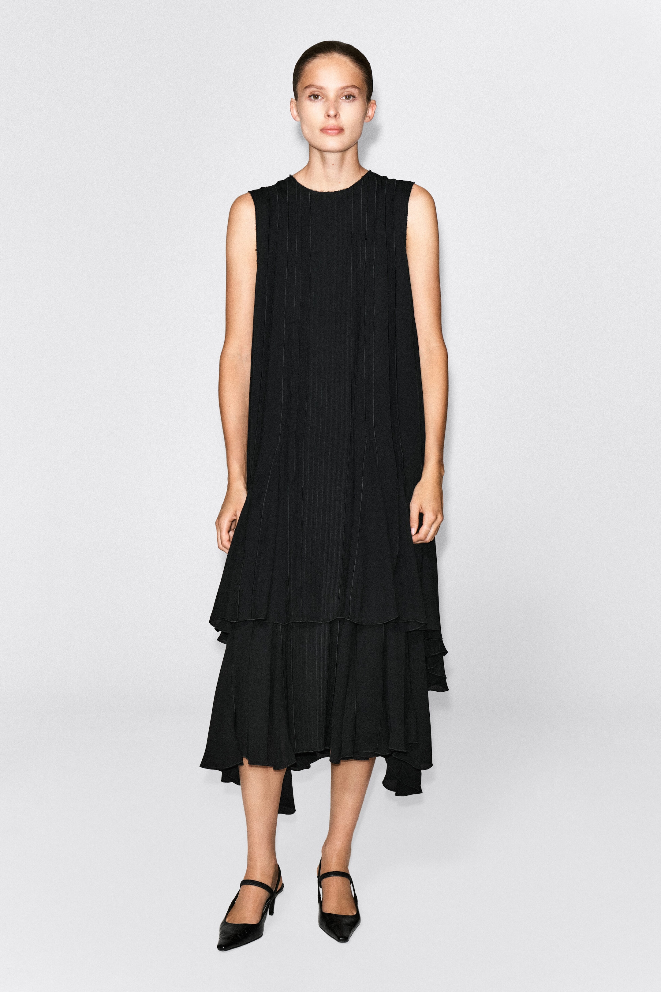 The model is wearing a black voluminous pleated midi dress, featuring a sleeveless design, a high neckline, and fine vertical pleats. The dress falls to a tiered, asymmetrical hem and is styled with black pointed-toe heels. The model is wearing a black voluminous pleated midi dress, featuring a sleeveless design, a high neckline, and fine vertical pleats. The dress falls to a tiered, asymmetrical hem and is styled with black pointed-toe heels.
