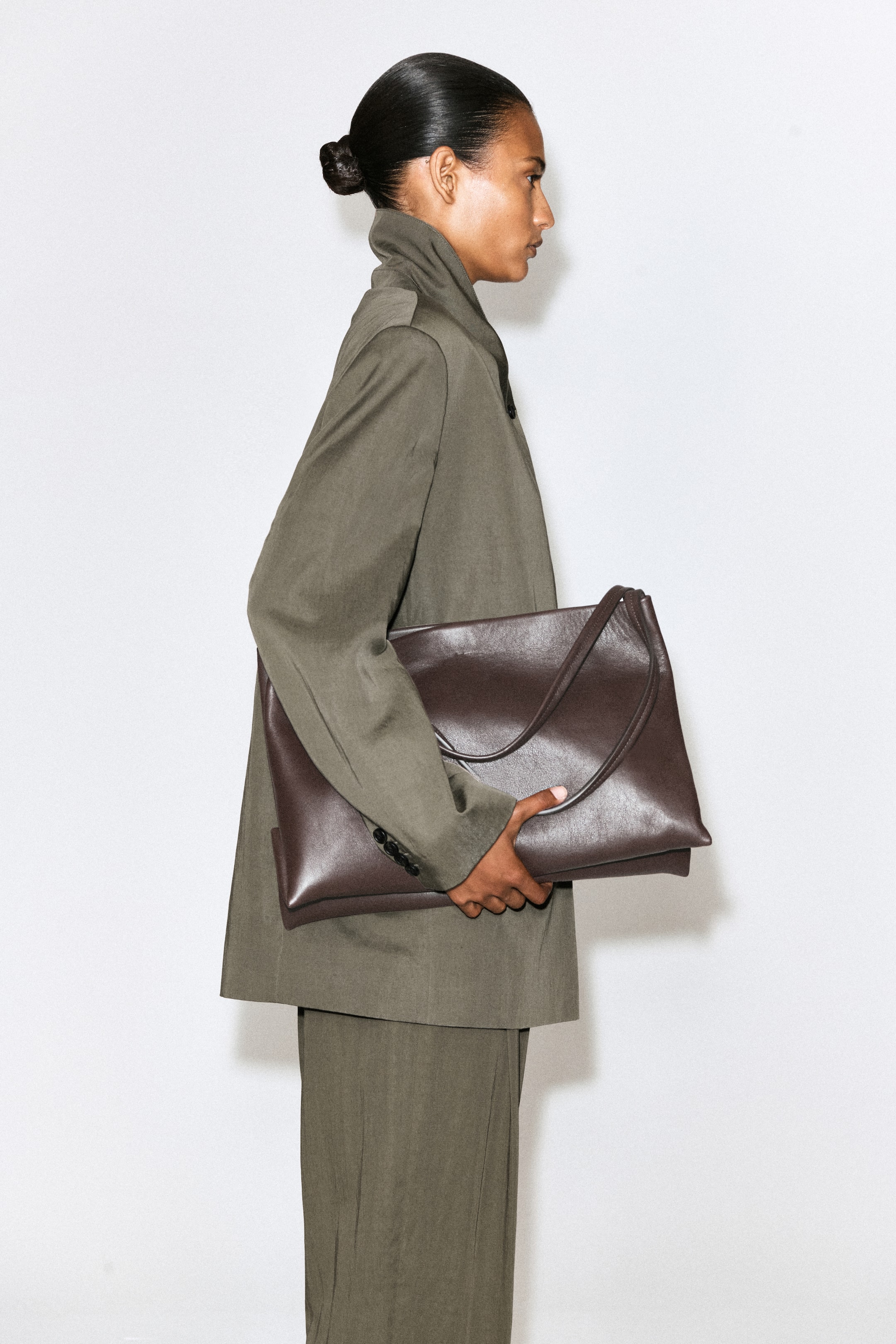 Größeres Bild anzeigen: Eine dunkelbraune Lederschultertasche mit weichem, rechteckigem Korpus und einem einzelnen Riemen wird vom Model gehalten. Das Model trägt die Tasche seitlich an den Körper angelegt und hält dabei ihren unteren Rand fest.
