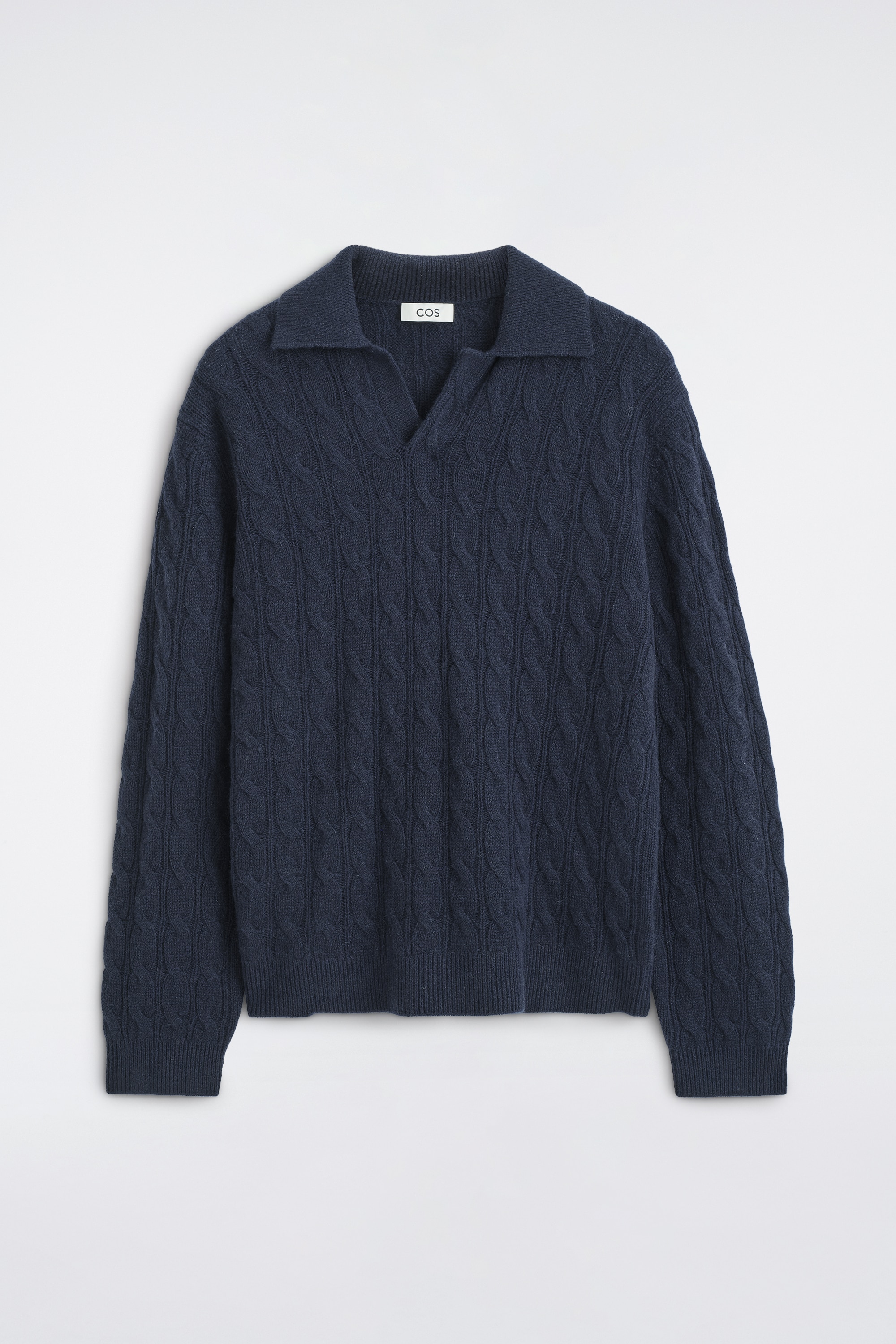 Il maglione polo in cashmere a trecce blu navy è indossato dalla modella, abbinato a una T-shirt bianca con girocollo e a pantaloni neri. Questo maglione a maniche lunghe presenta un colletto polo aperto, una lavorazione a trecce all-over e bordi a costine su orlo e polsini.